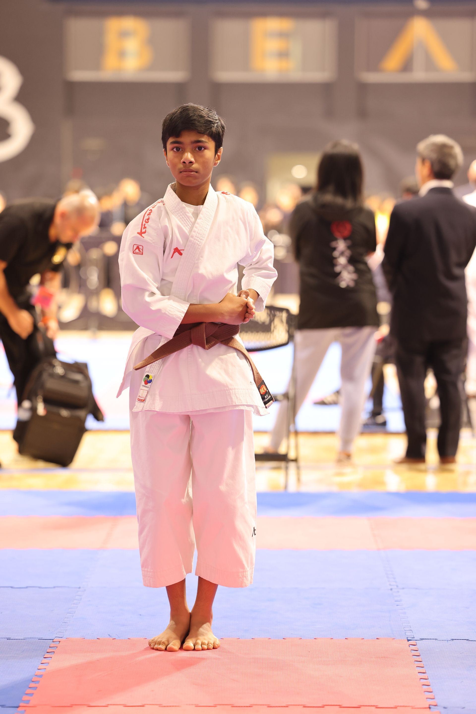 Martial artist in a white uniform stands on a red mat at an indoor competition, arms crossed.
