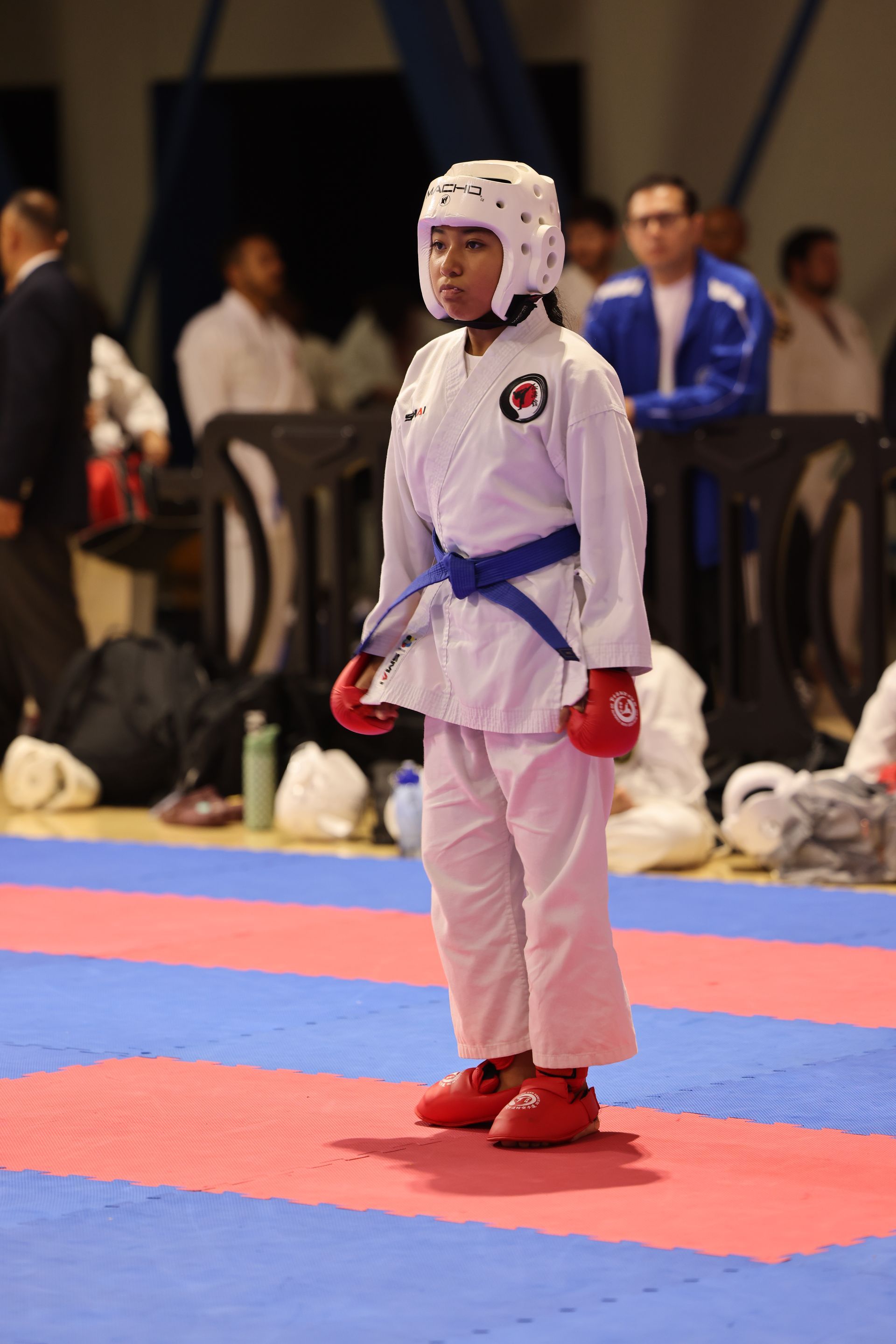 Young martial artist in white gi and blue belt standing on a red-and-blue mat, wearing protective headgear and gloves