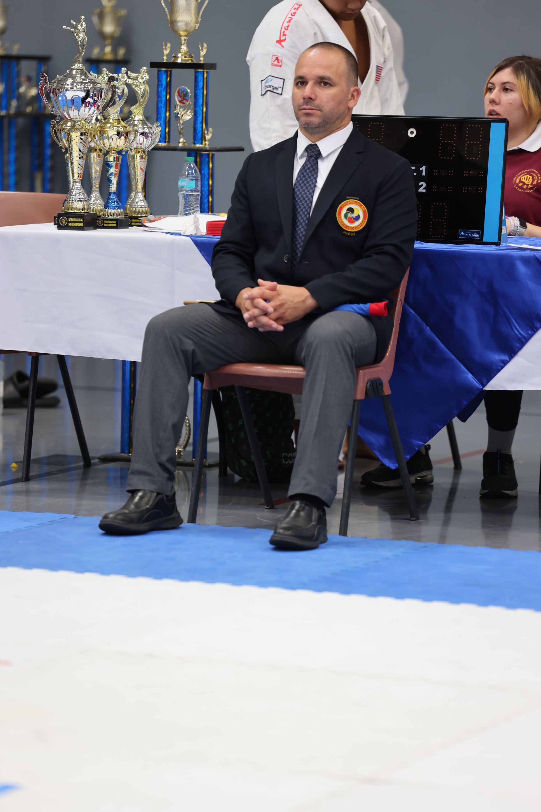 Man in suit and tie seated at table, watching a competition. Trophies and blue/white tablecloth present.