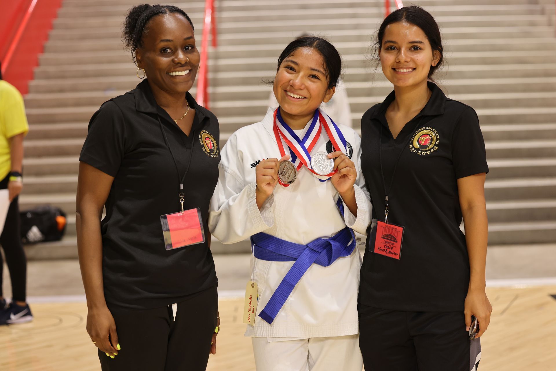 Three people smiling on a gym floor, one in a white martial arts uniform with a medal and blue belt.