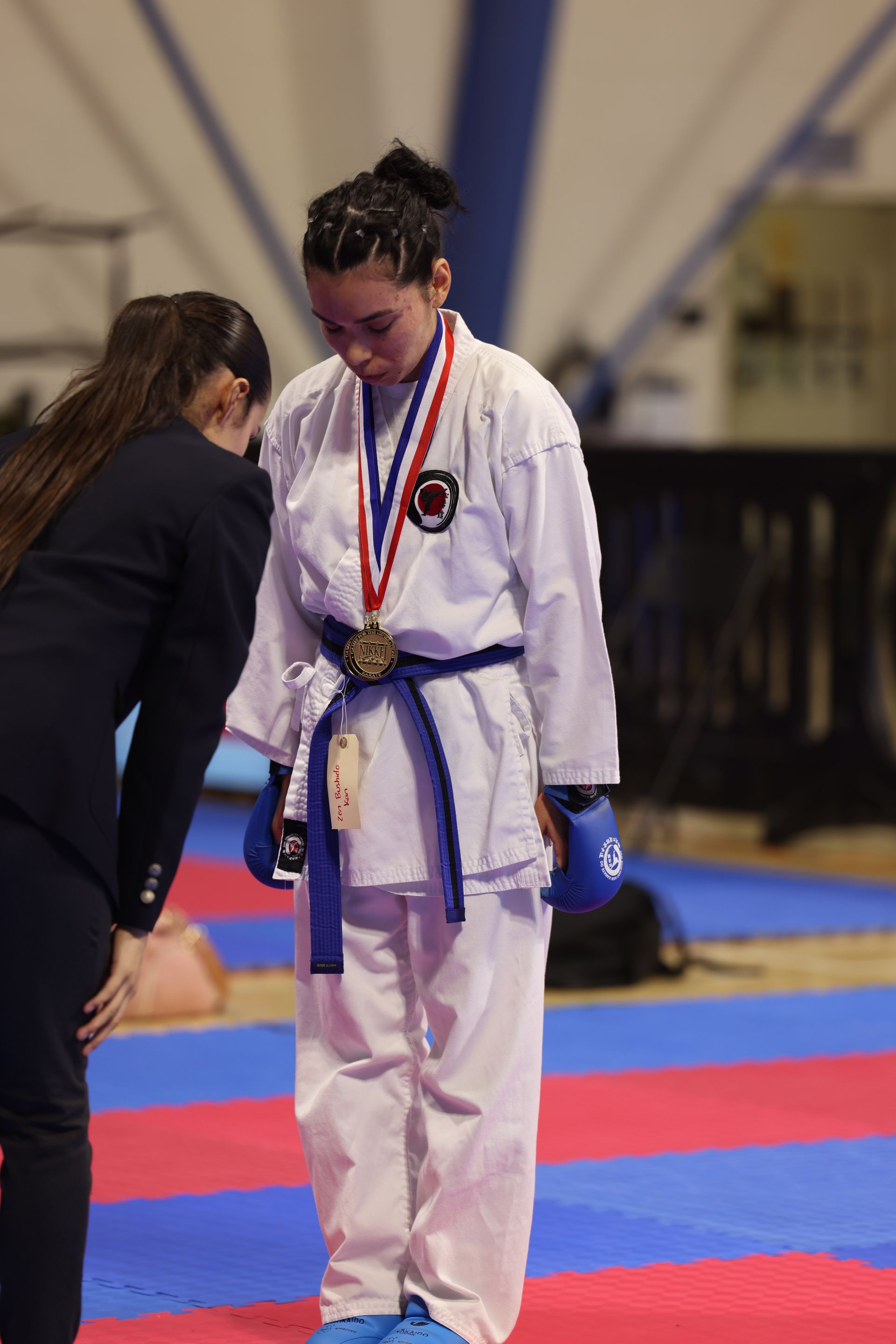Karate competitor in white gi with medals stands on a mat as an official speaks beside them