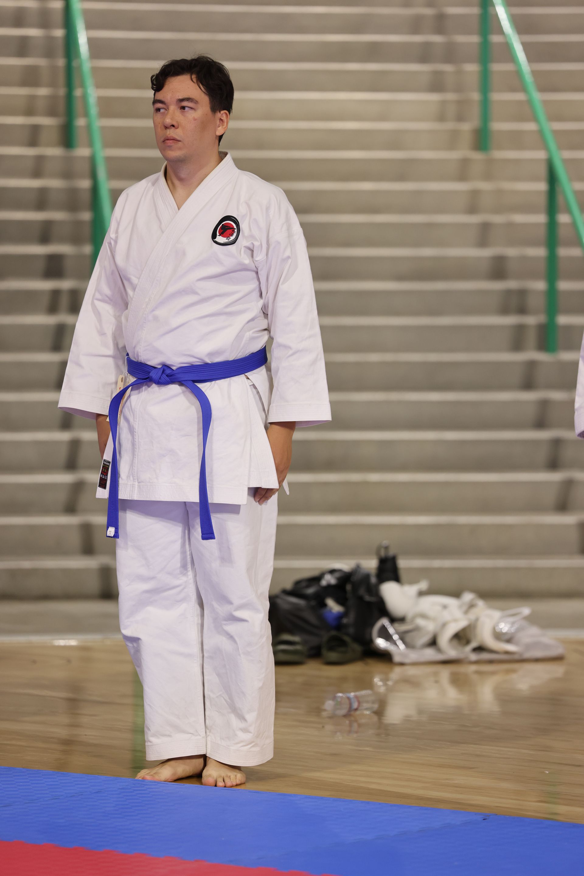 Martial artist in white gi with blue belt standing on a mat in a gymnasium