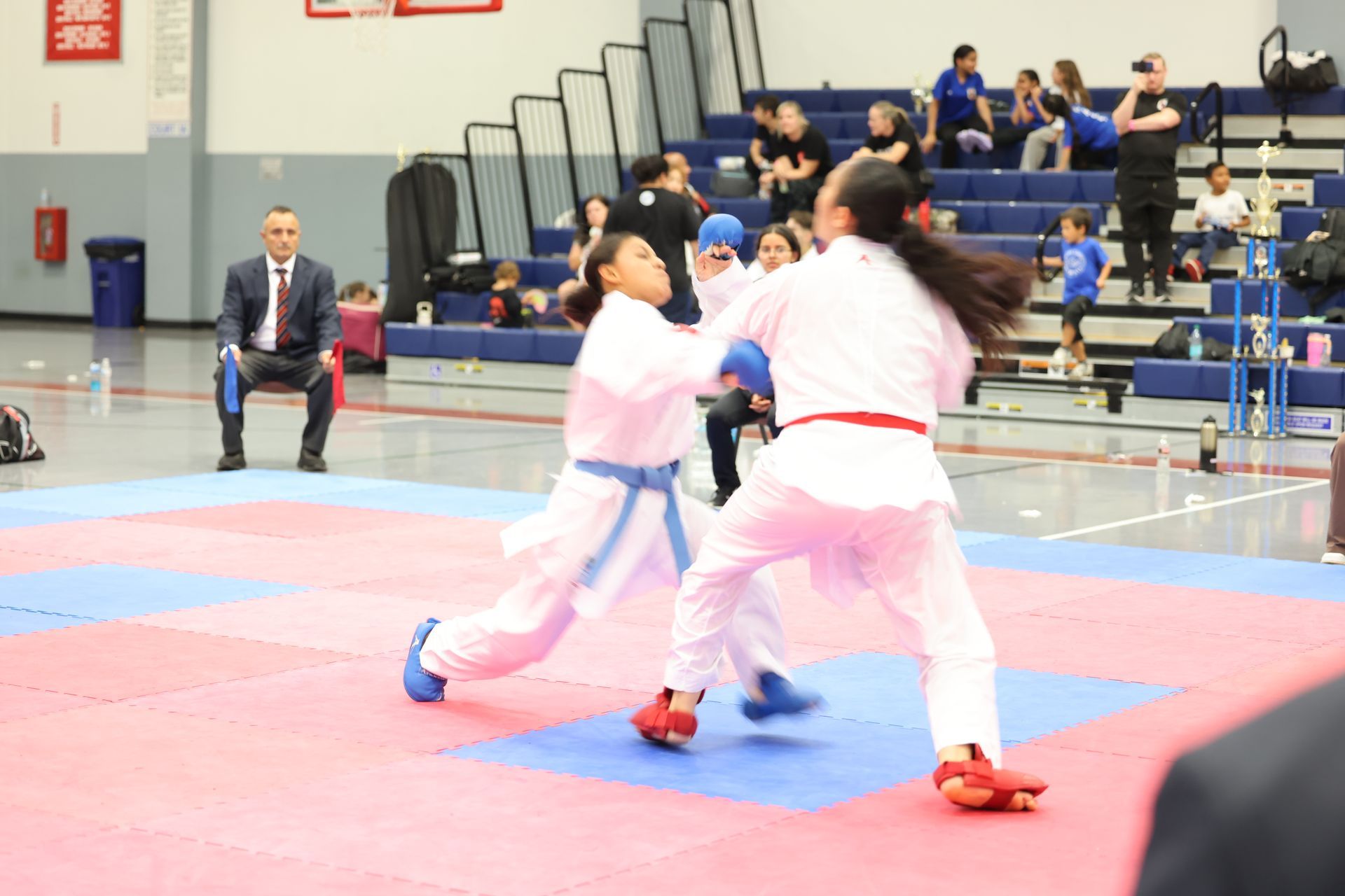 Two karate athletes in white gis with blue and red belts, in a match, with a judge and spectators.