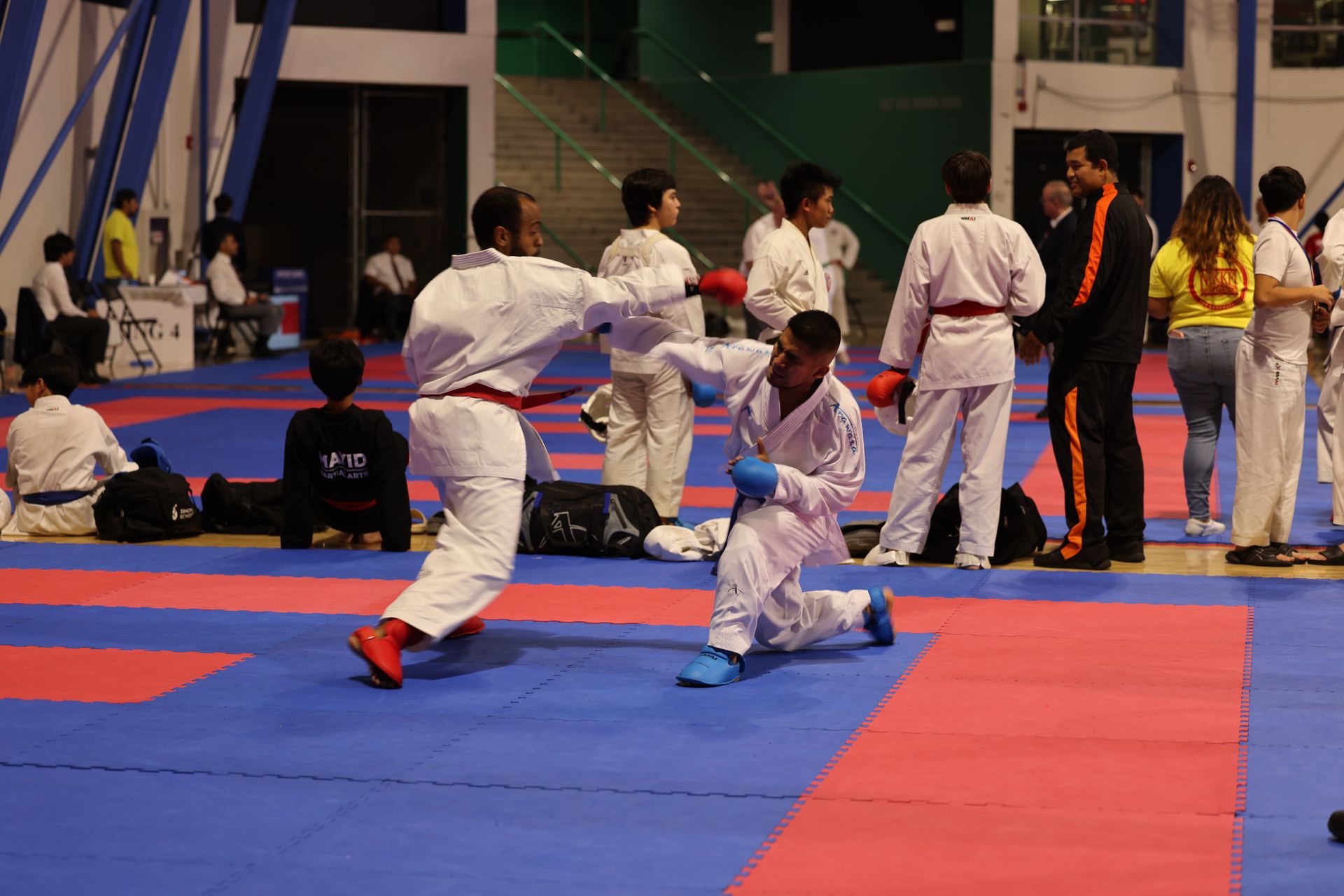 Martial arts sparring match on blue and red mats, with two athletes in white uniforms facing off.