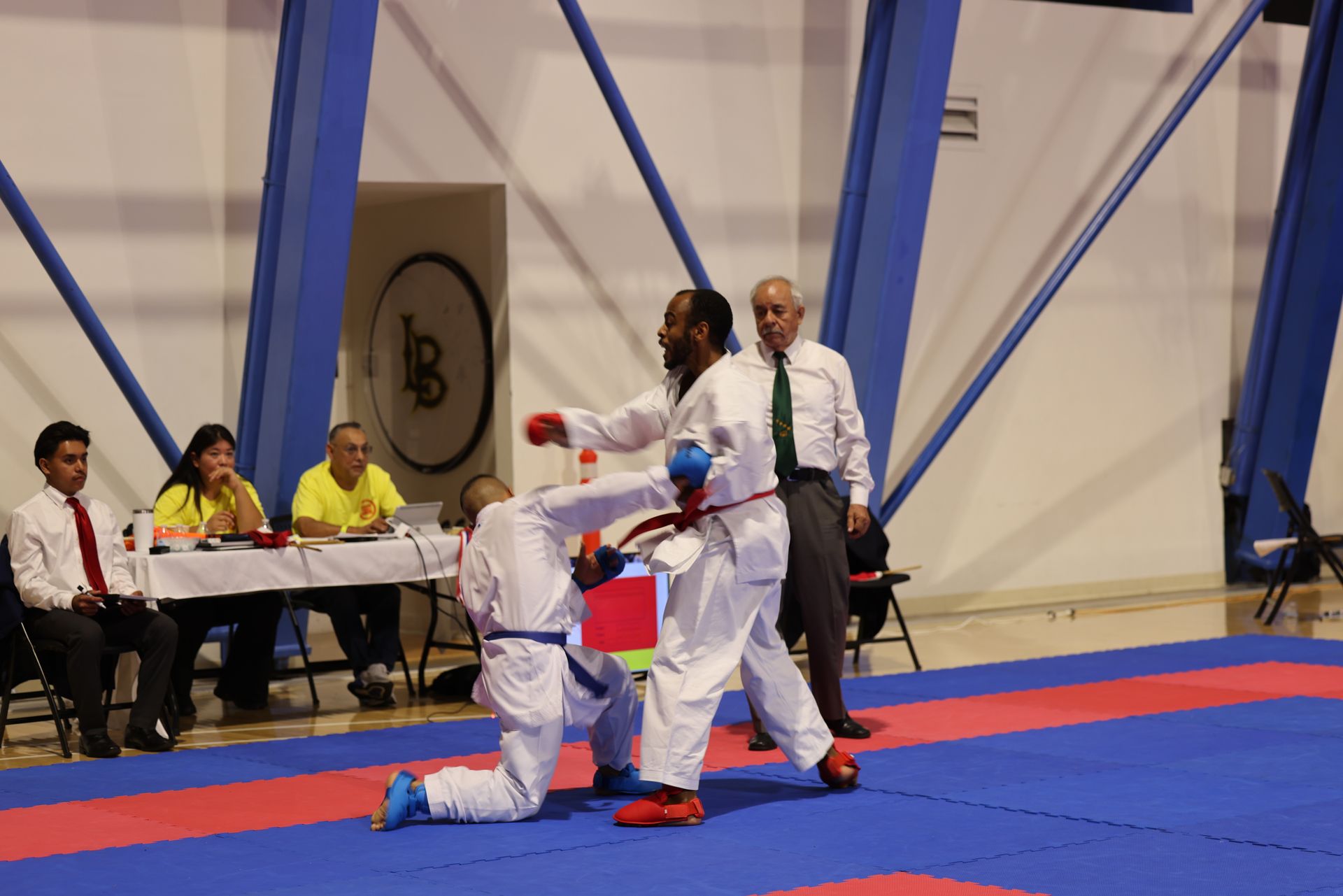 Taekwondo match with two competitors and a referee on a blue mat near judges’ table