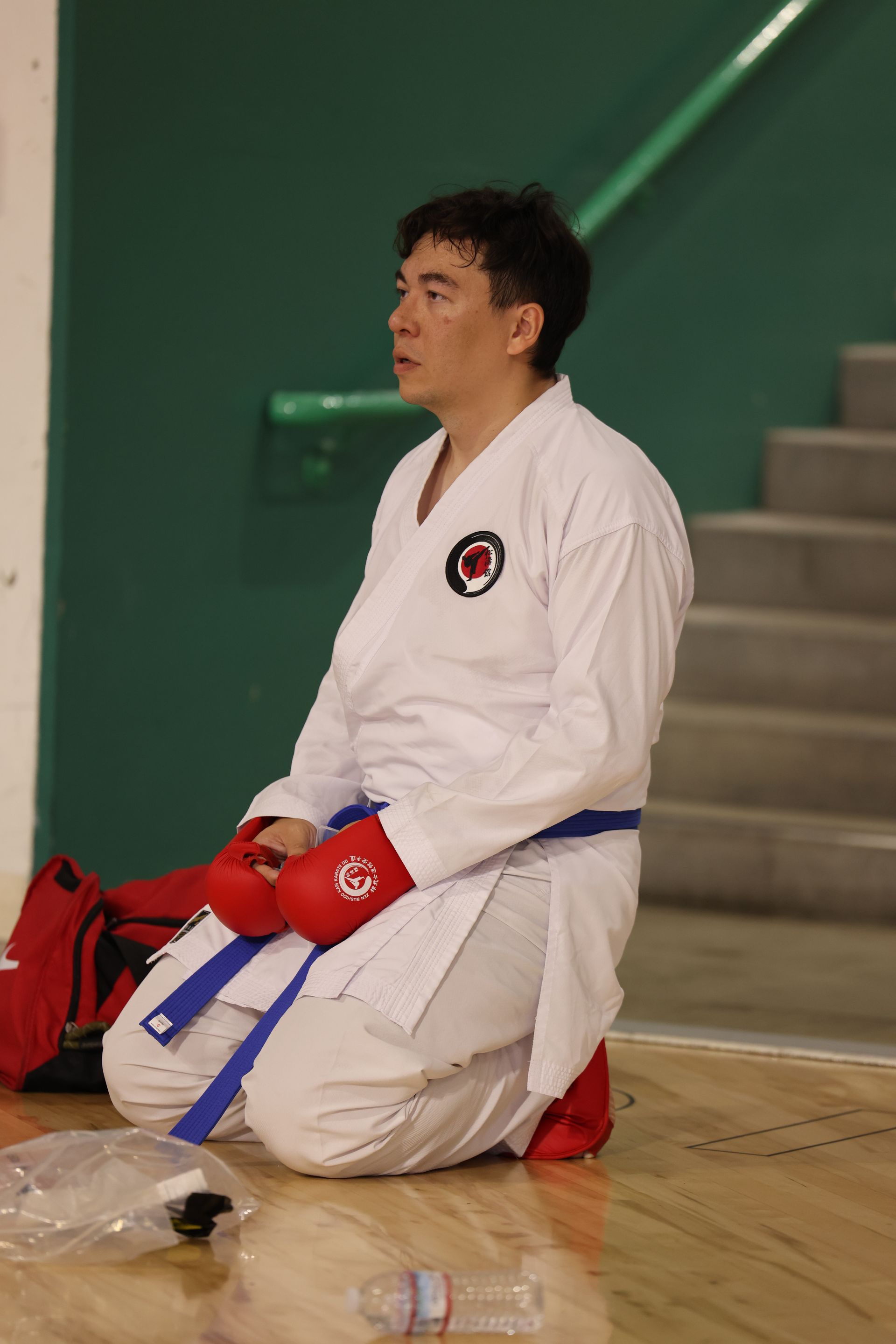 Martial artist in white gi and red gloves kneels on a gym floor beside a red bag and blue belt.