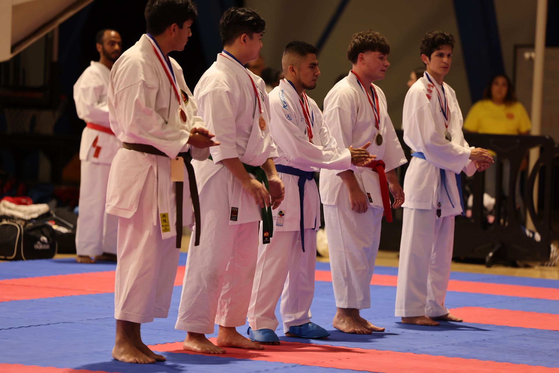 Five karate competitors in white gis stand on a blue and red mat during a competition.