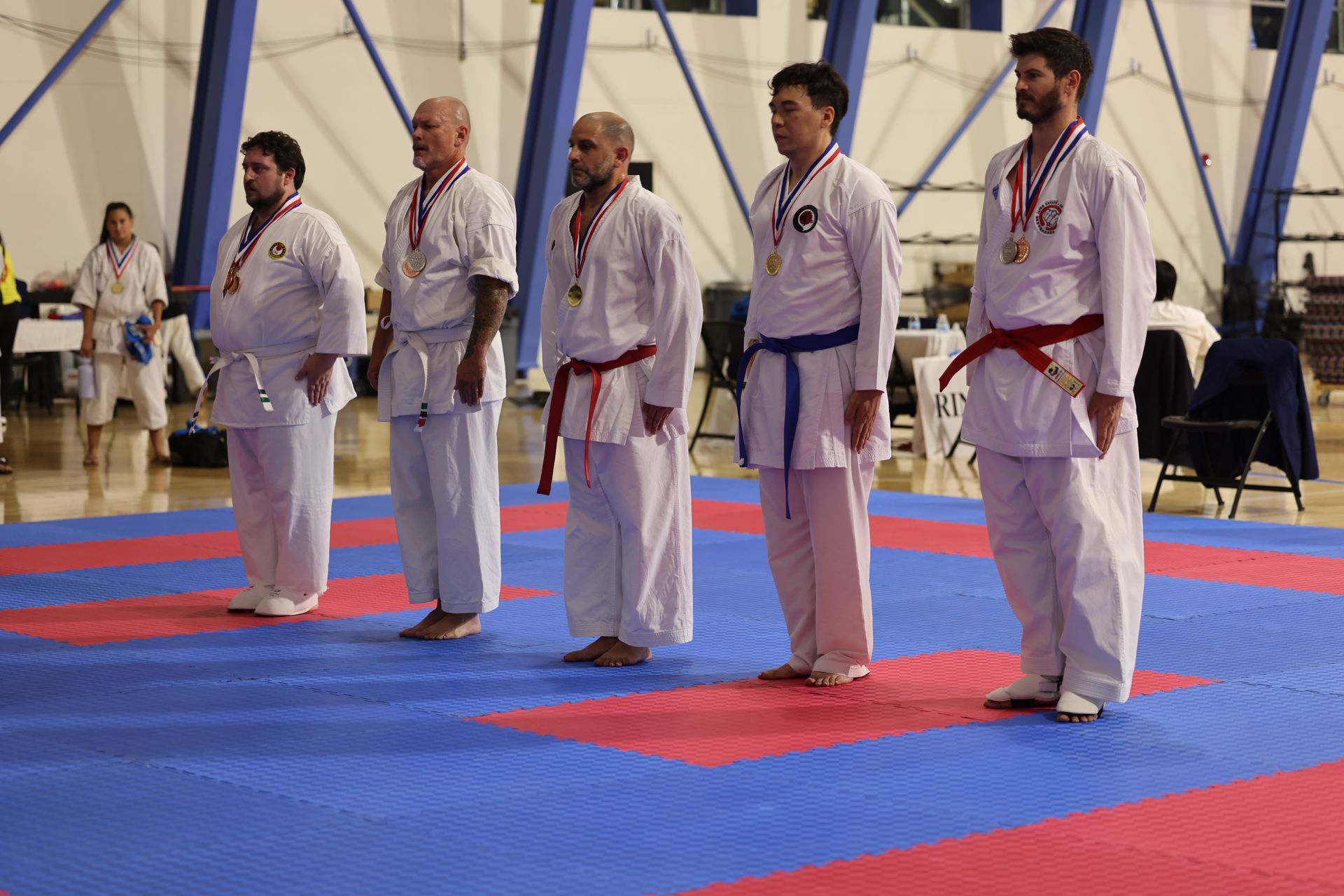 Five people in white martial arts uniforms stand on blue and red mats in an indoor arena.