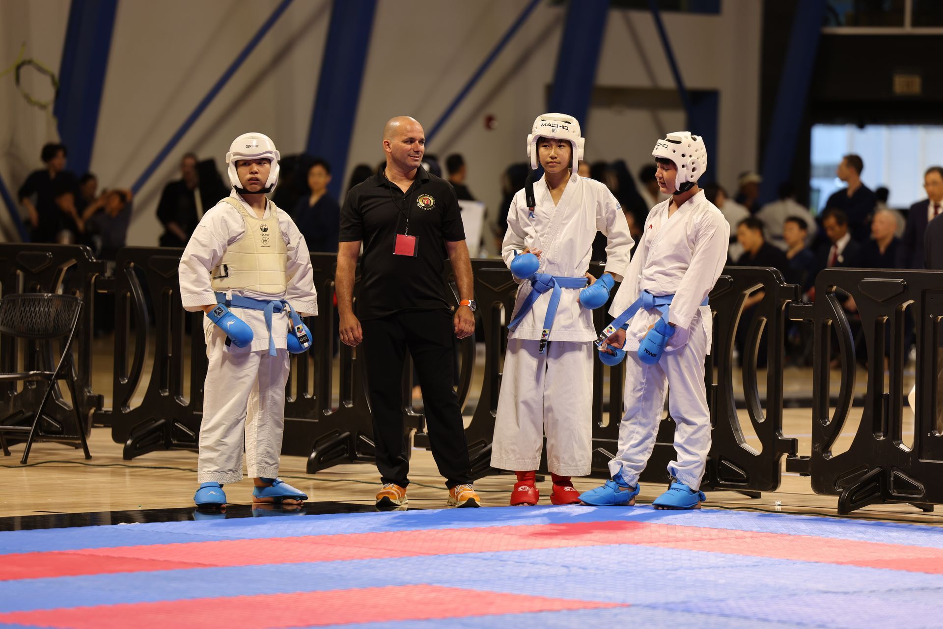 Three martial artists in white uniforms and blue gloves stand on a mat with a referee in a sports arena