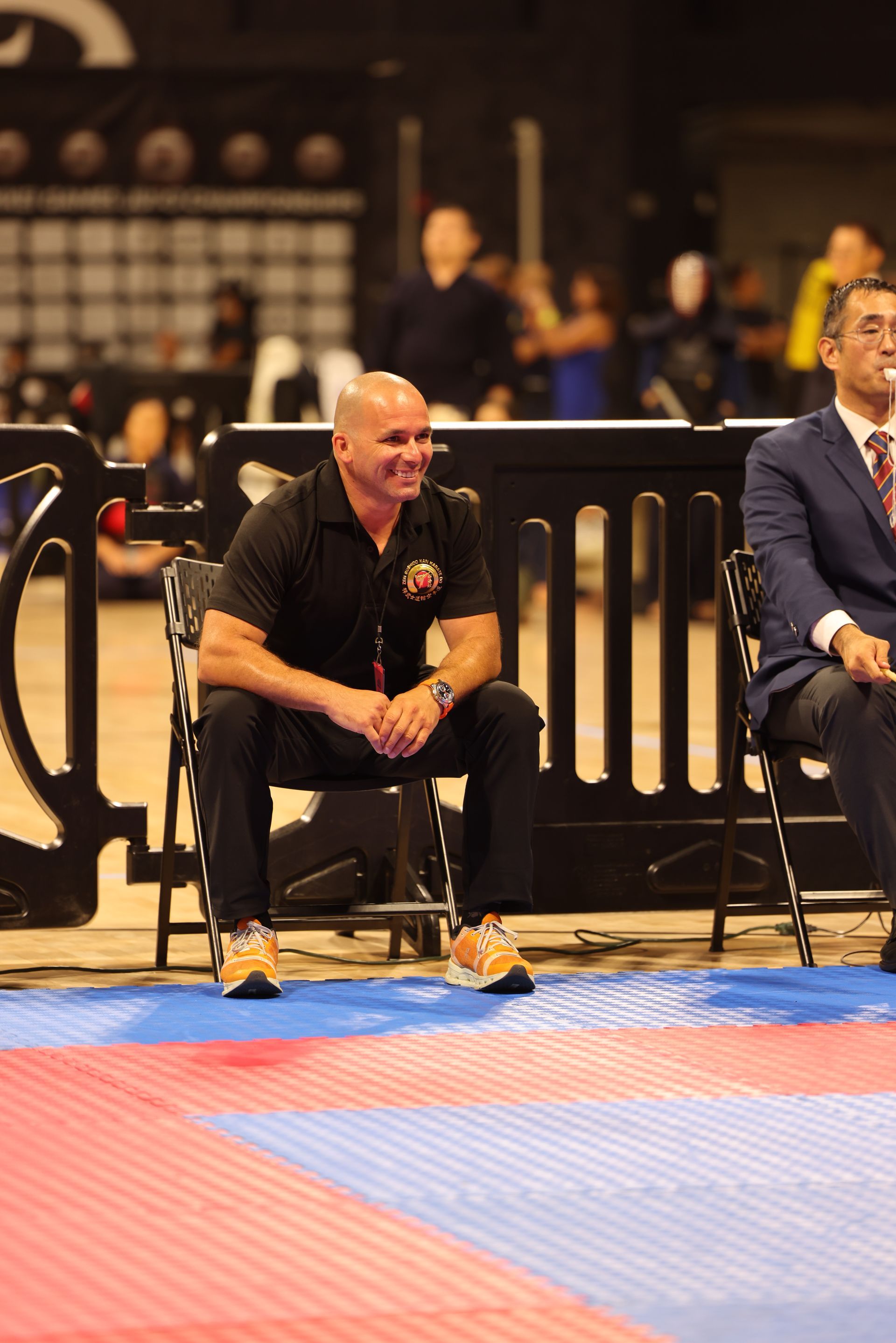 Man crouching beside a mat at an indoor sports event, wearing black with orange shoes.