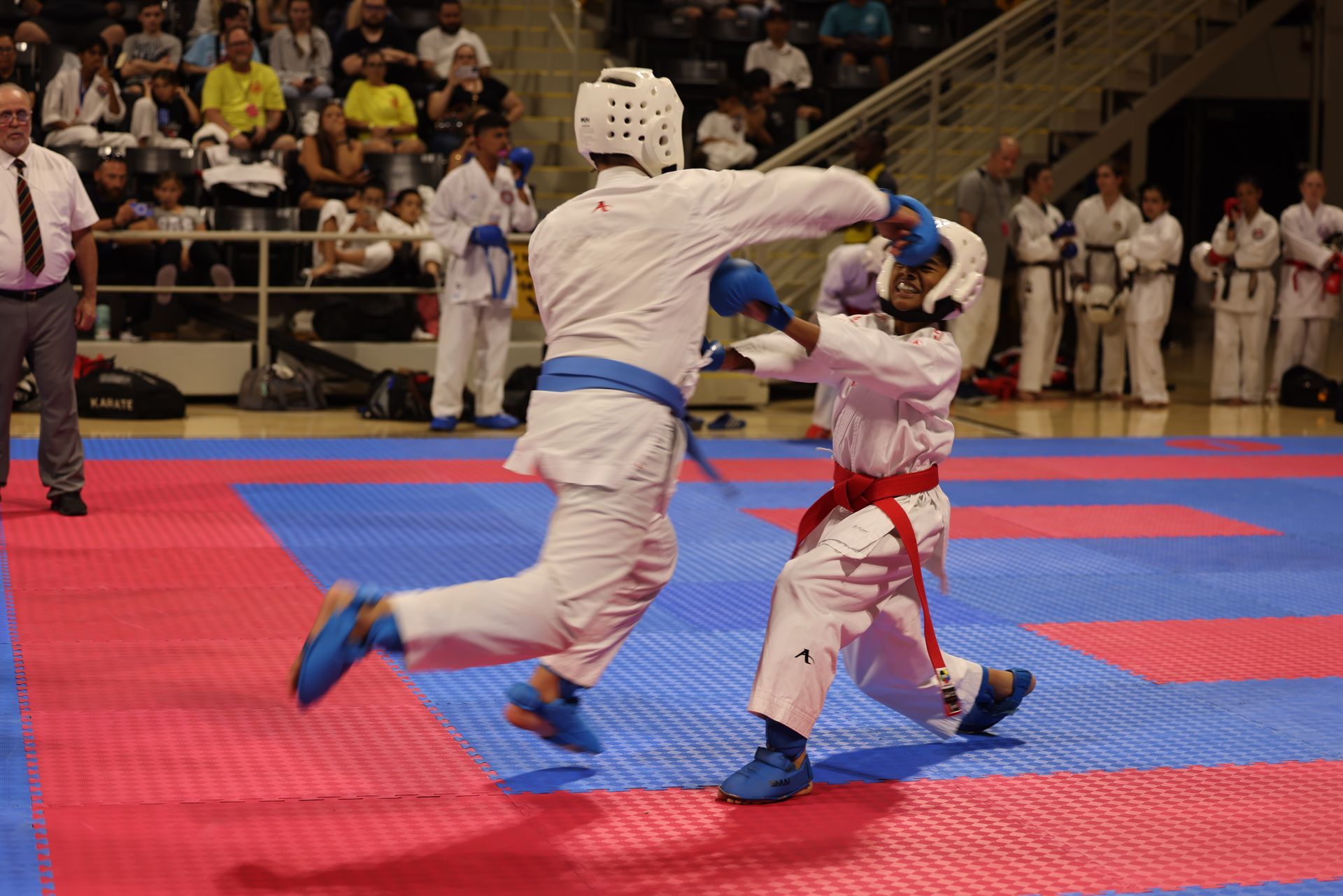 Two martial artists sparring on a red-and-blue mat in a tournament gym, one kicking midair.