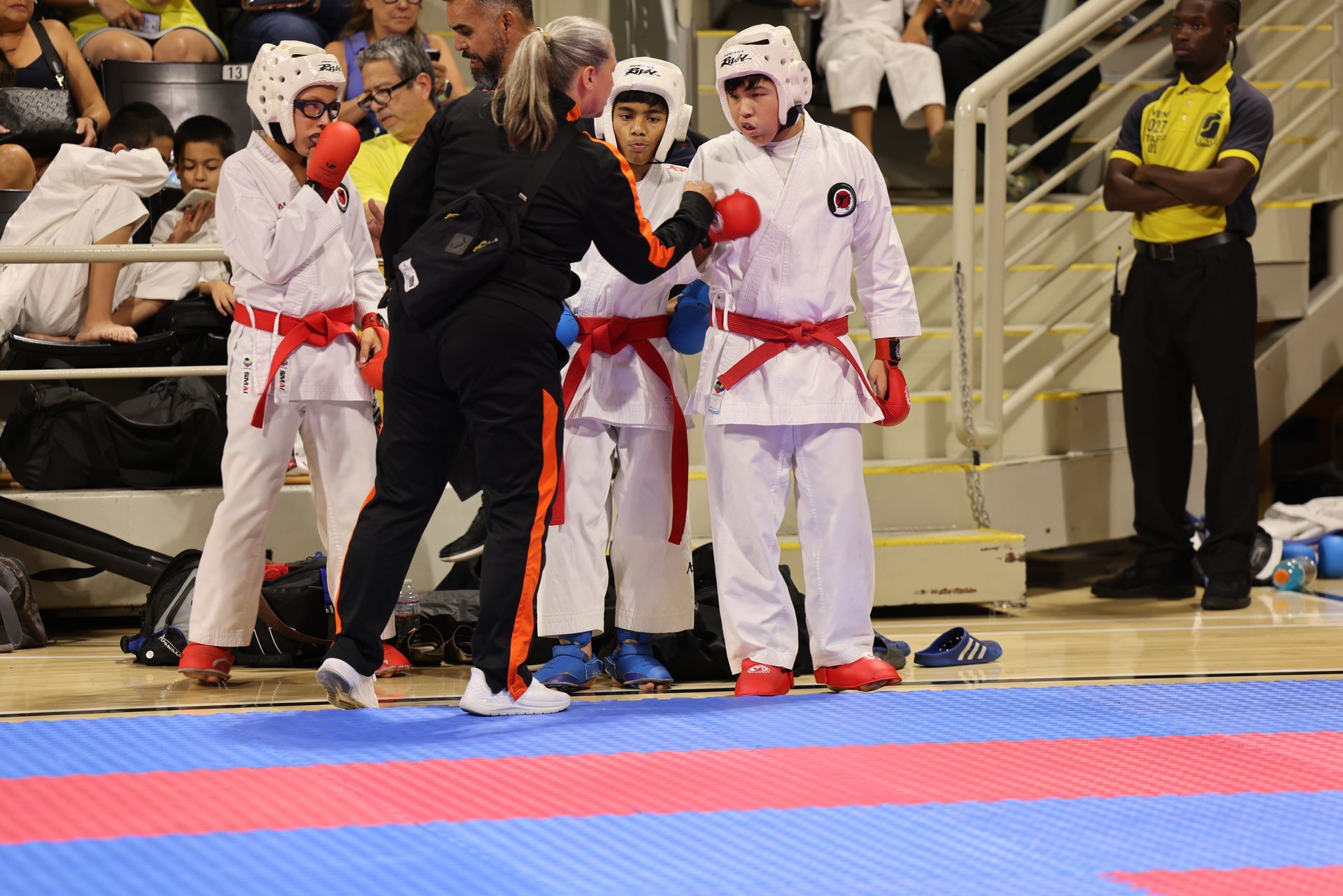 Karate competitors in white uniforms with red belts stand on a blue and red mat during a match.