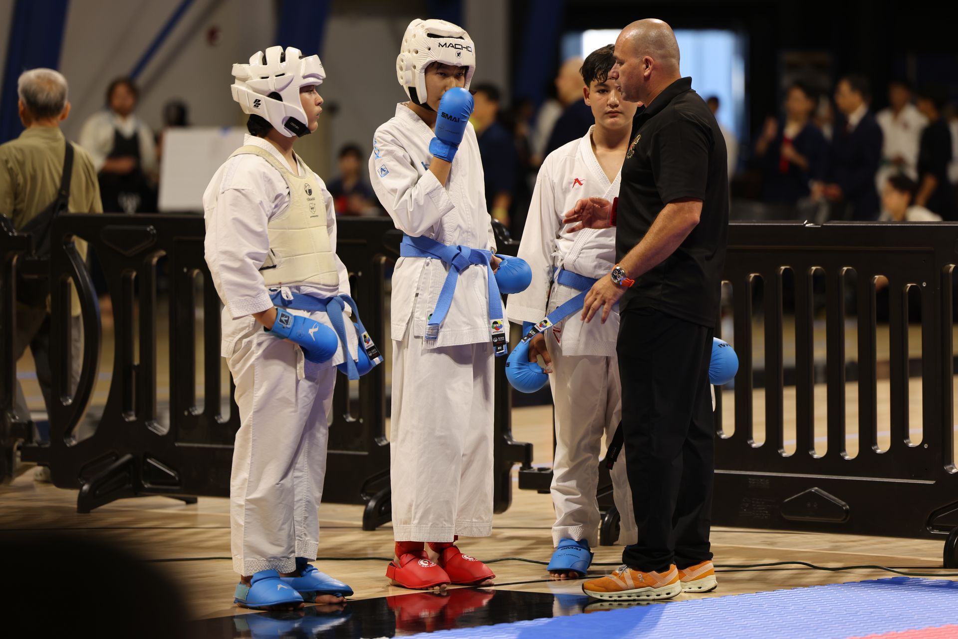 Three martial artists in white uniforms and blue gloves talk with a referee before a match.