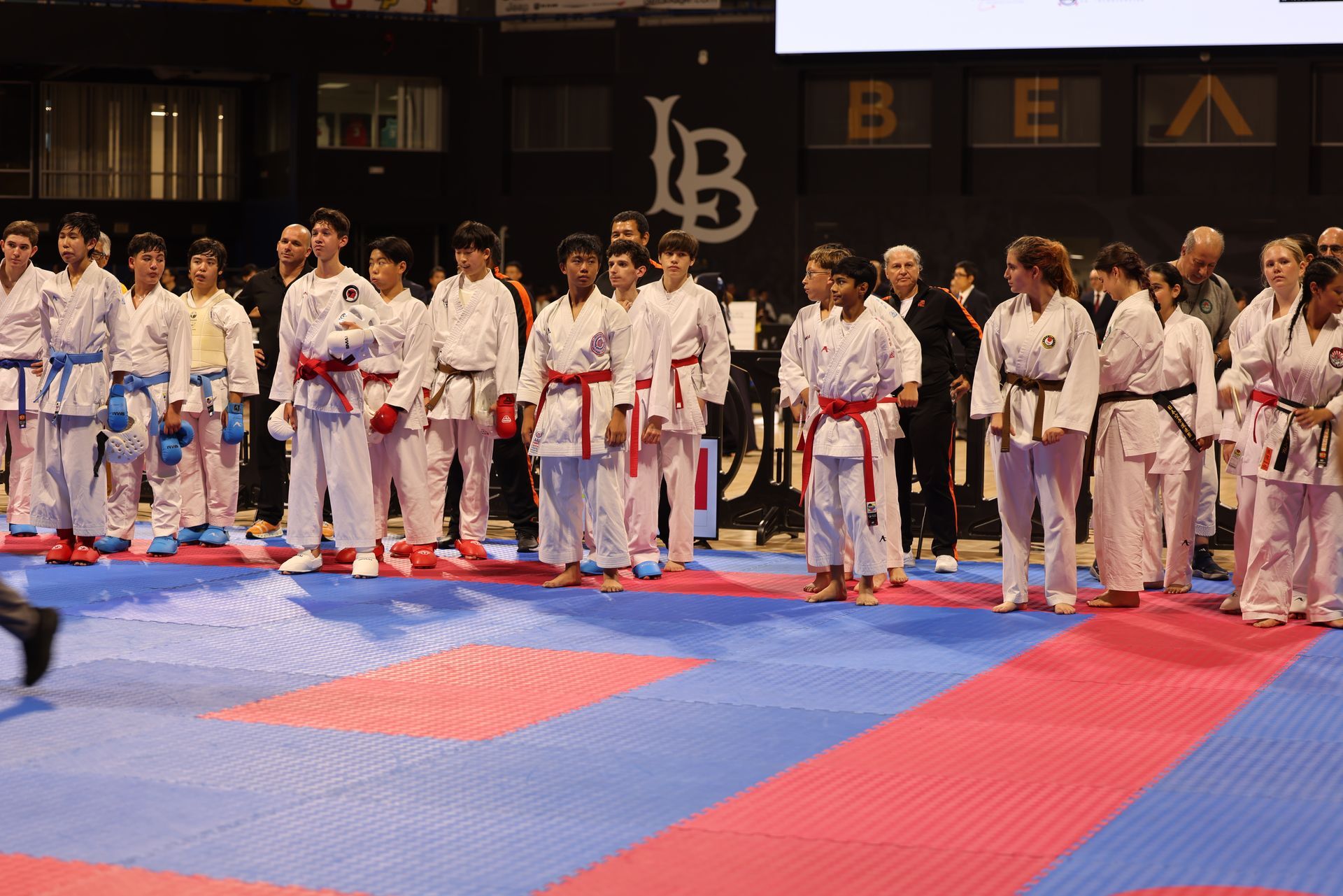 Martial arts competitors in white uniforms lined up on red and blue mats in an indoor arena