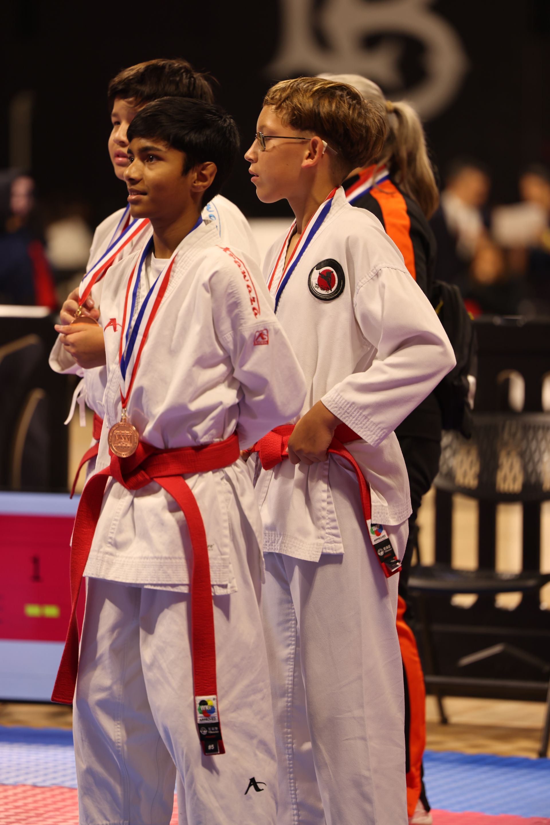 Two martial artists in white uniforms with red belts standing on a competition mat