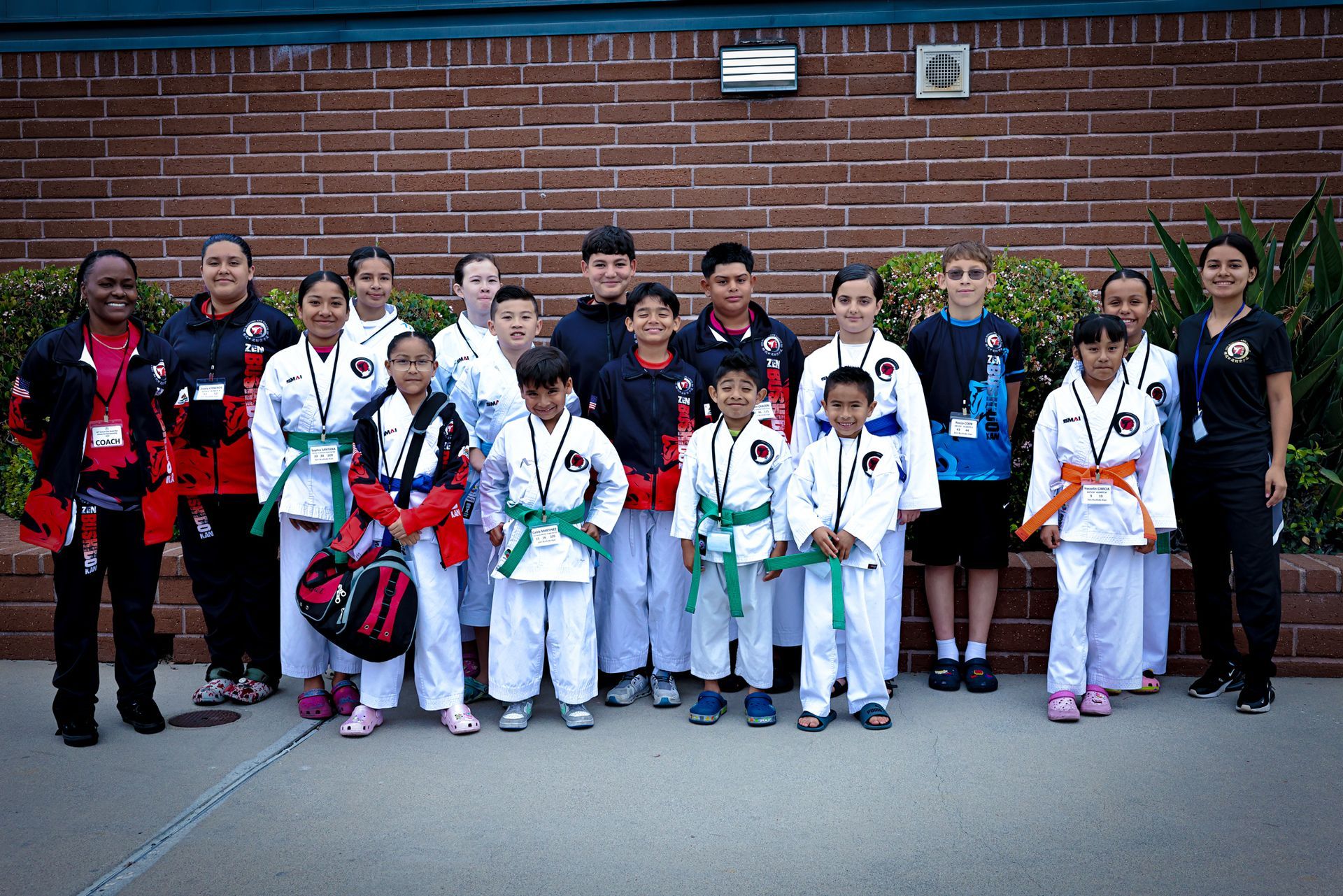 A group of youth martial arts students in white uniforms and a few instructors stand against a brick wall.