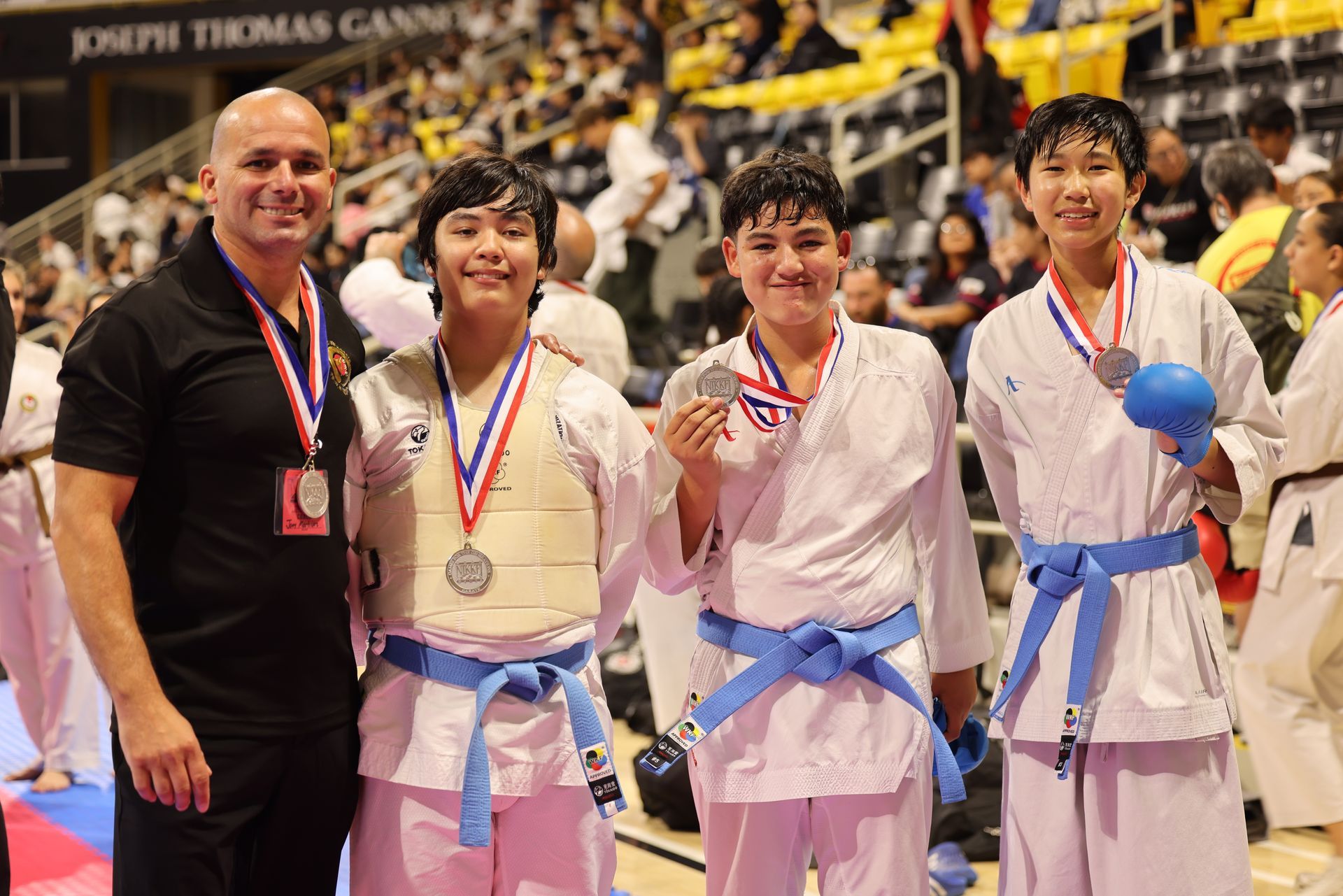 Three medal-winning martial artists pose with a coach at a tournament, holding blue sparring gear.