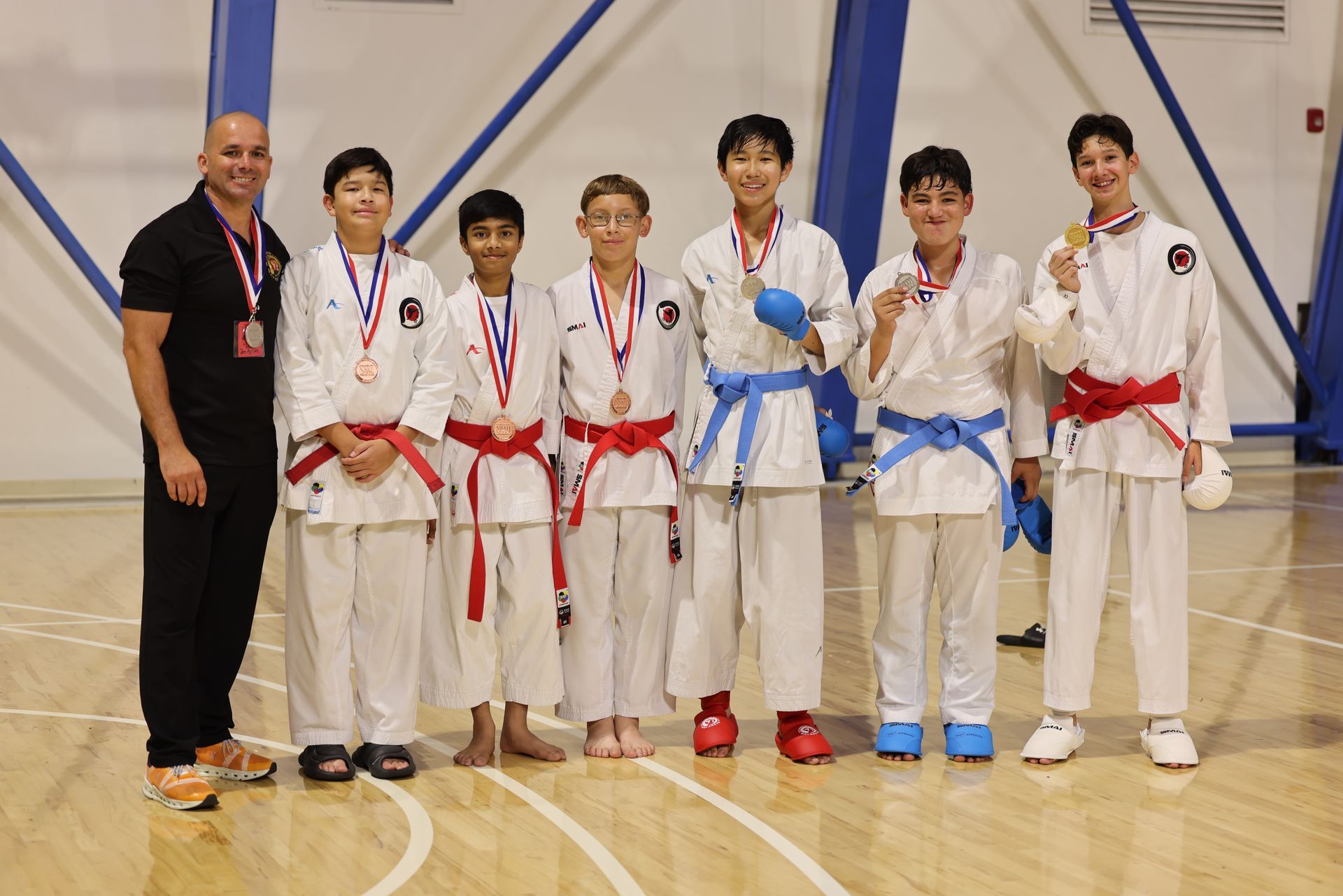 Karate team with medals and trophies posing in a gym, wearing white gis with red and blue belts.