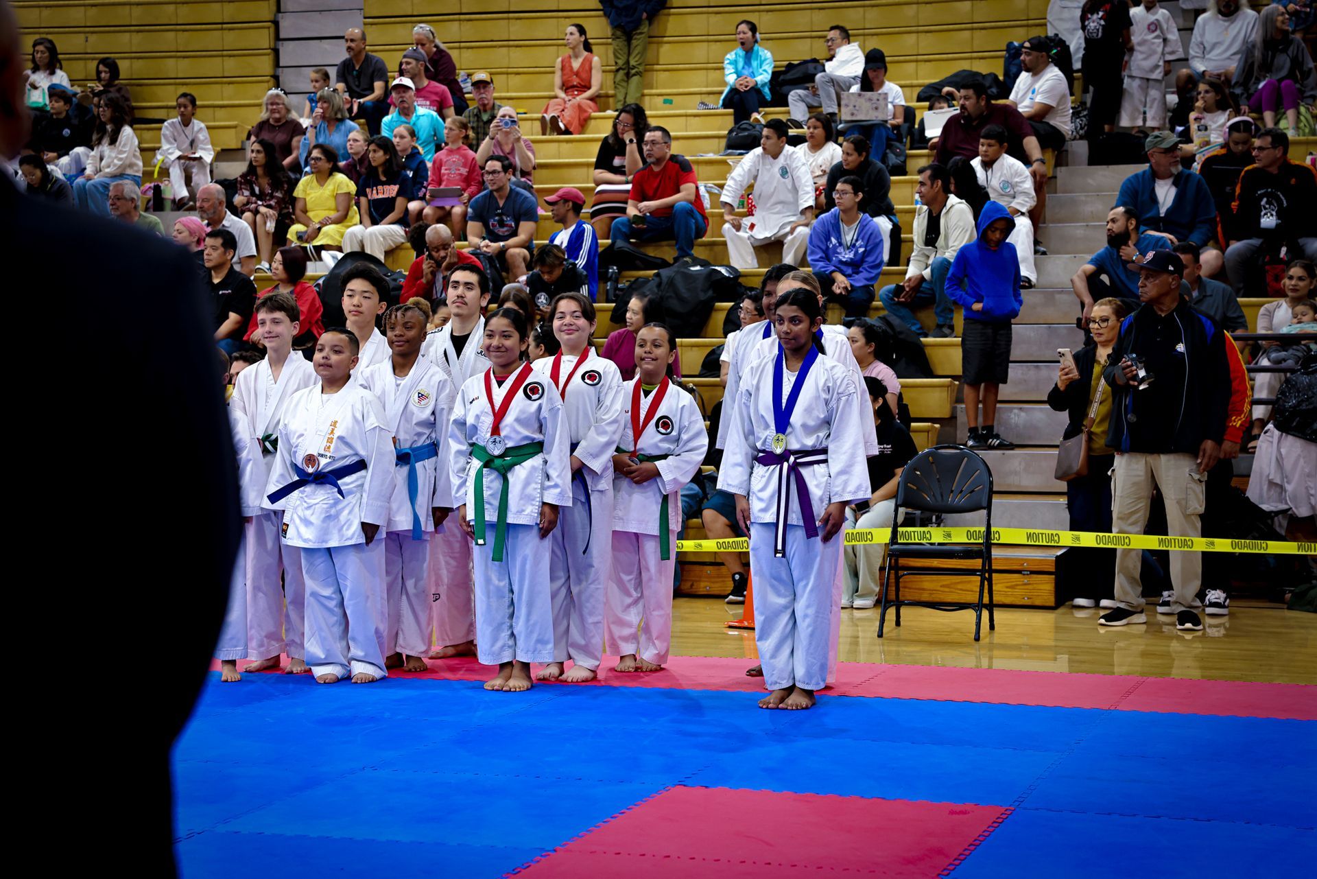 A row of students in white karate uniforms with colored belts stand on blue and red mats during a martial arts tournament.