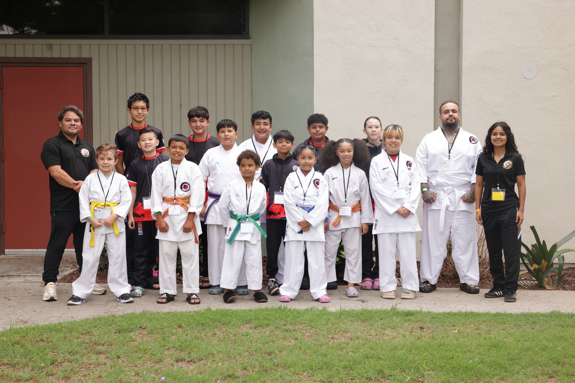 Group of martial arts students and instructors posing in white uniforms and belts outside a building.