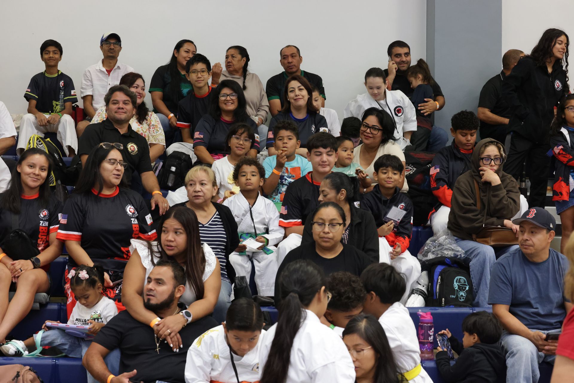 Crowd seated in bleachers at an indoor event, many wearing black shirts with red accents.