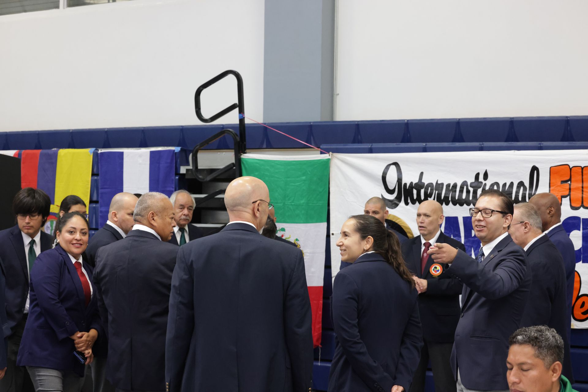 People in suits mingling at an indoor event beside a banner and colorful flags