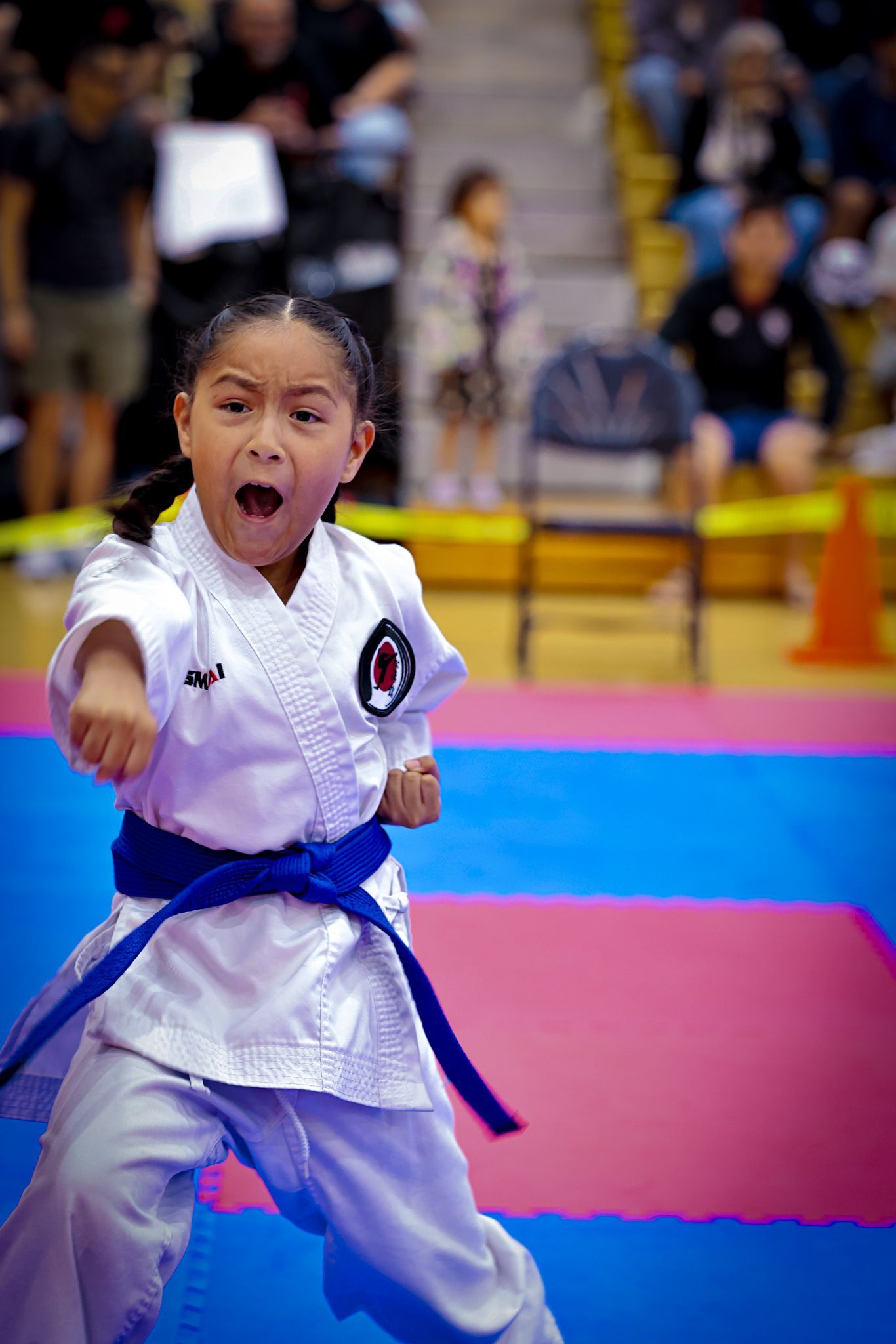 A child in a white karate uniform with a blue belt performs a punch with a focused expression on a blue and pink mat.