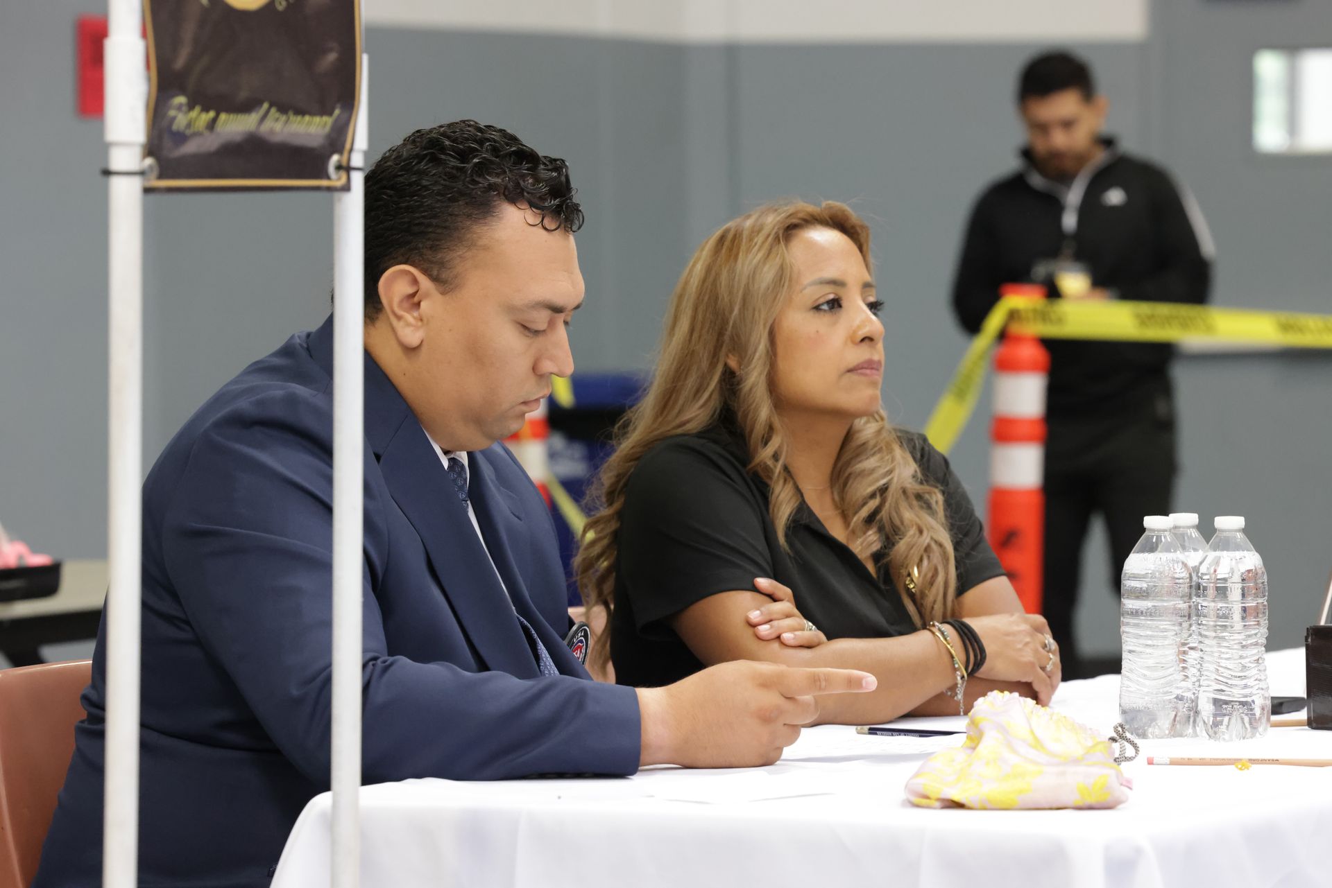 Two people seated at a table, taking notes during a formal meeting with water bottles and caution tape in the background