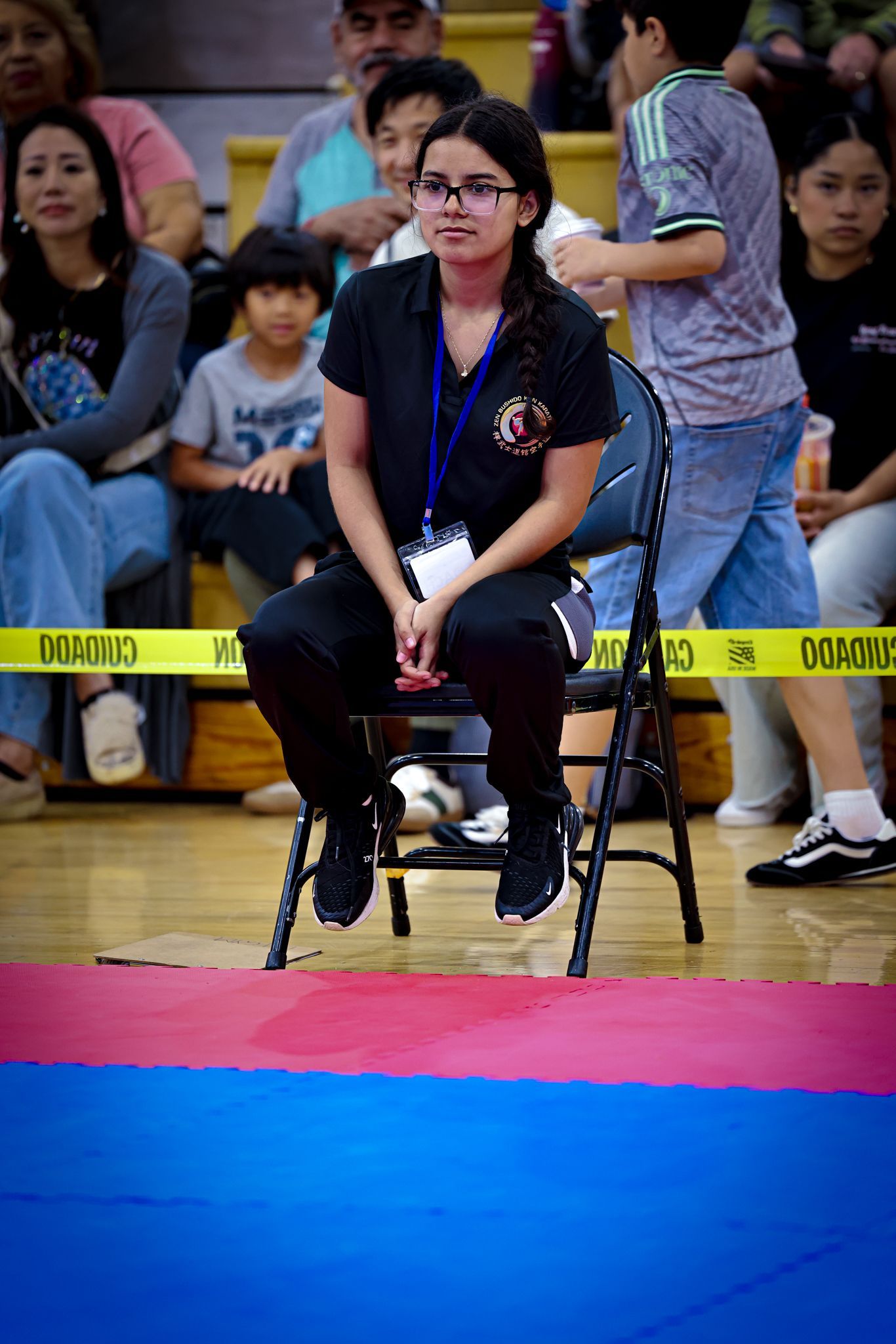 A person with glasses sits on a folding chair in an indoor gym, behind a yellow caution tape, watching a floor mat.