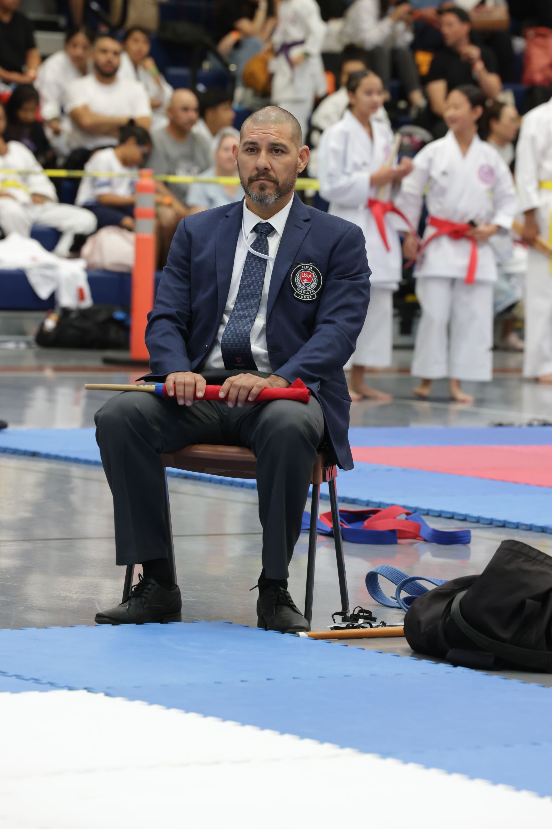 Man in a suit sits by a karate mat, holding red hand wraps during a martial arts match