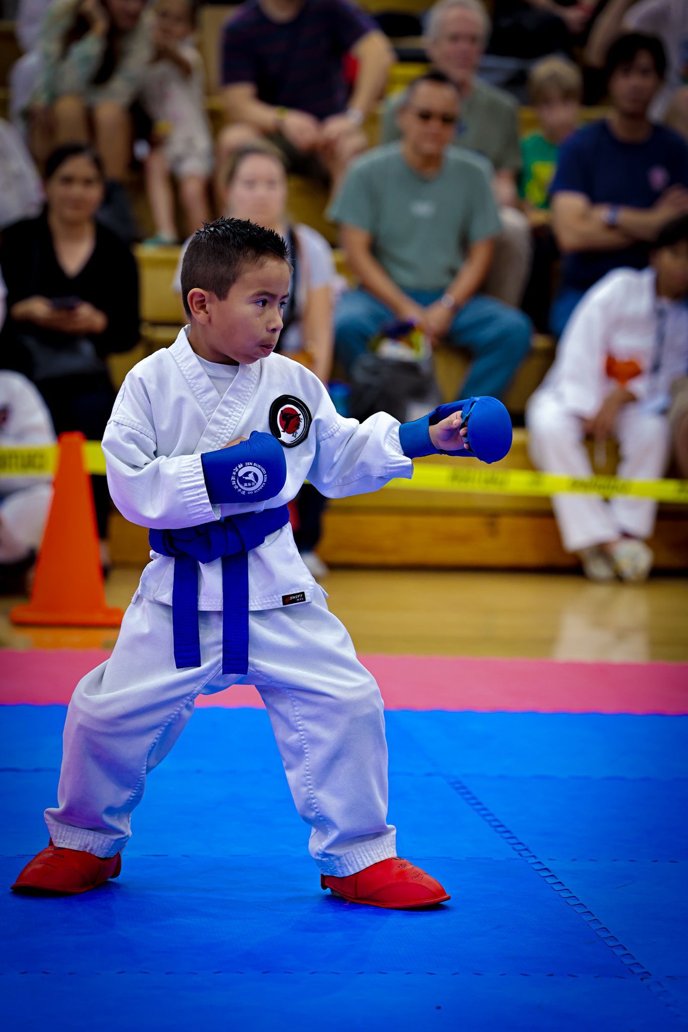 A child in a white karate uniform with a blue belt and gloves performs a punch on a blue mat in a gymnasium.
