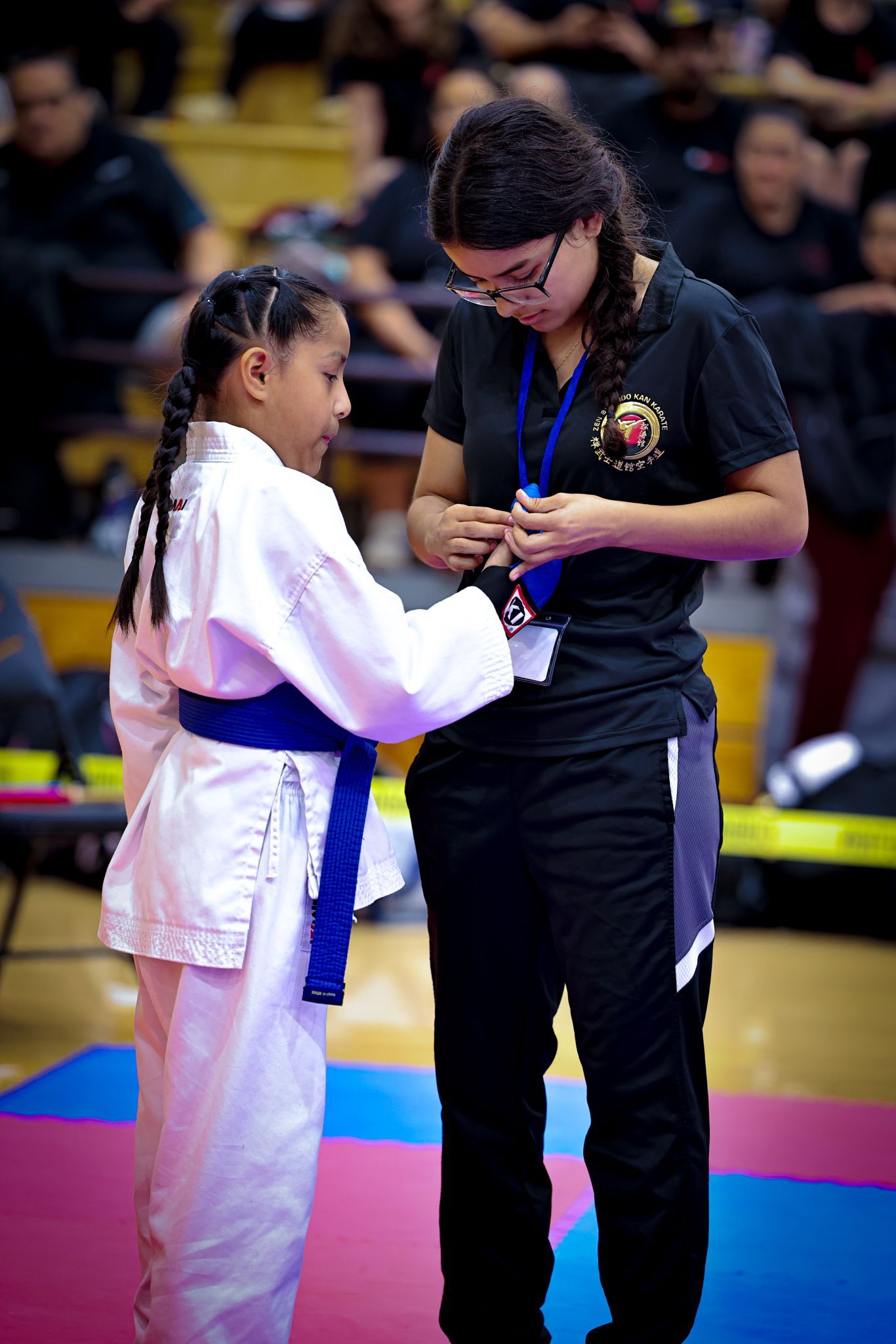 A girl in a white karate uniform with a blue belt receives a medal from a person in a black polo shirt in a gym.