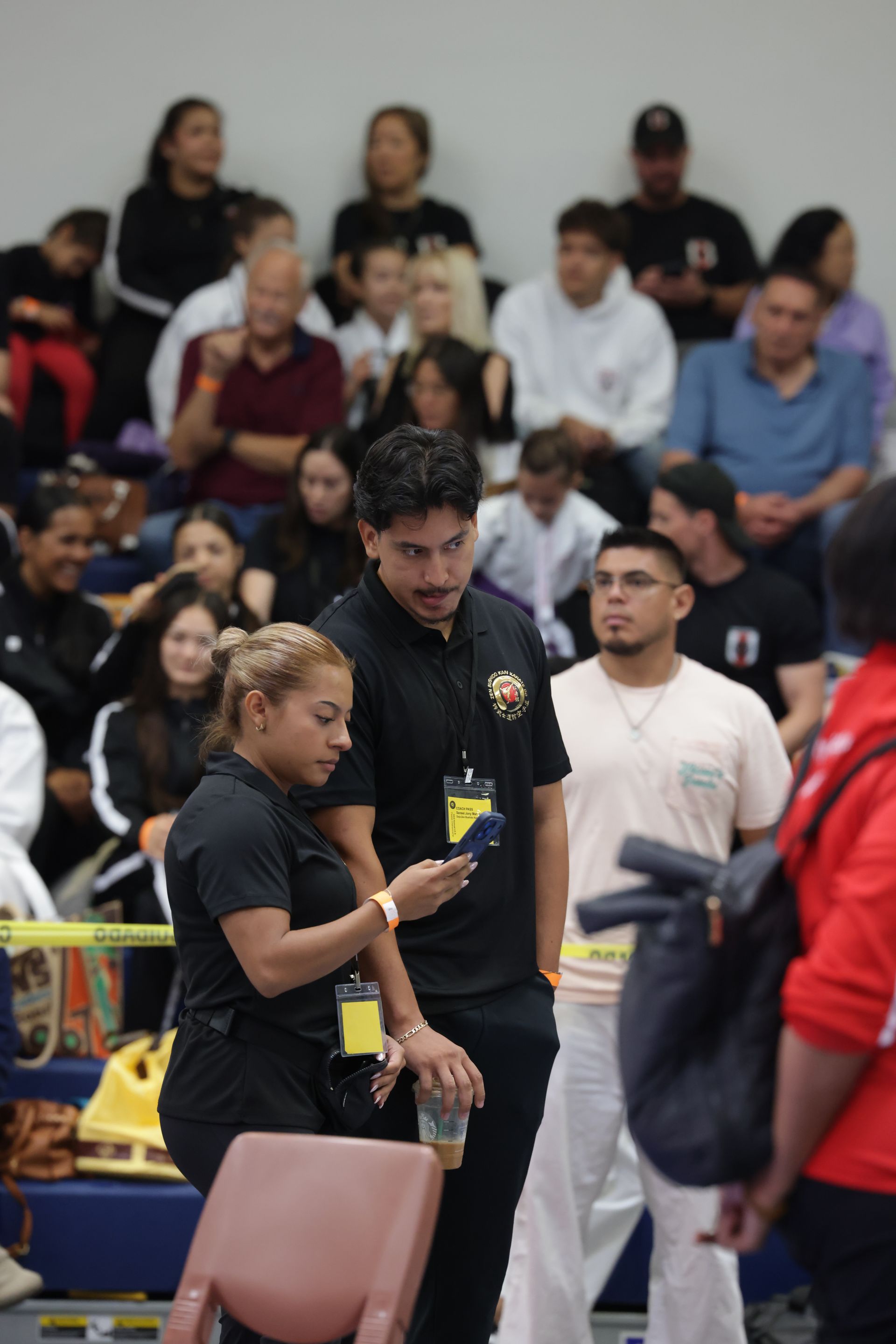 Crowd of people standing in an indoor arena, with two staff members in black shirts in the foreground.