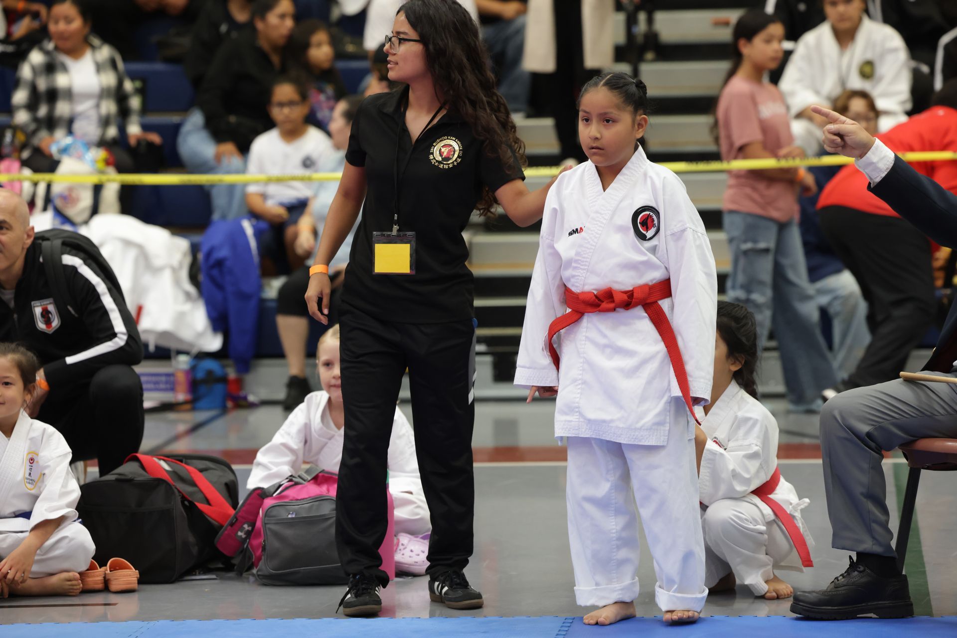 Martial arts competitor in white gi with red belt standing at a tournament beside a referee