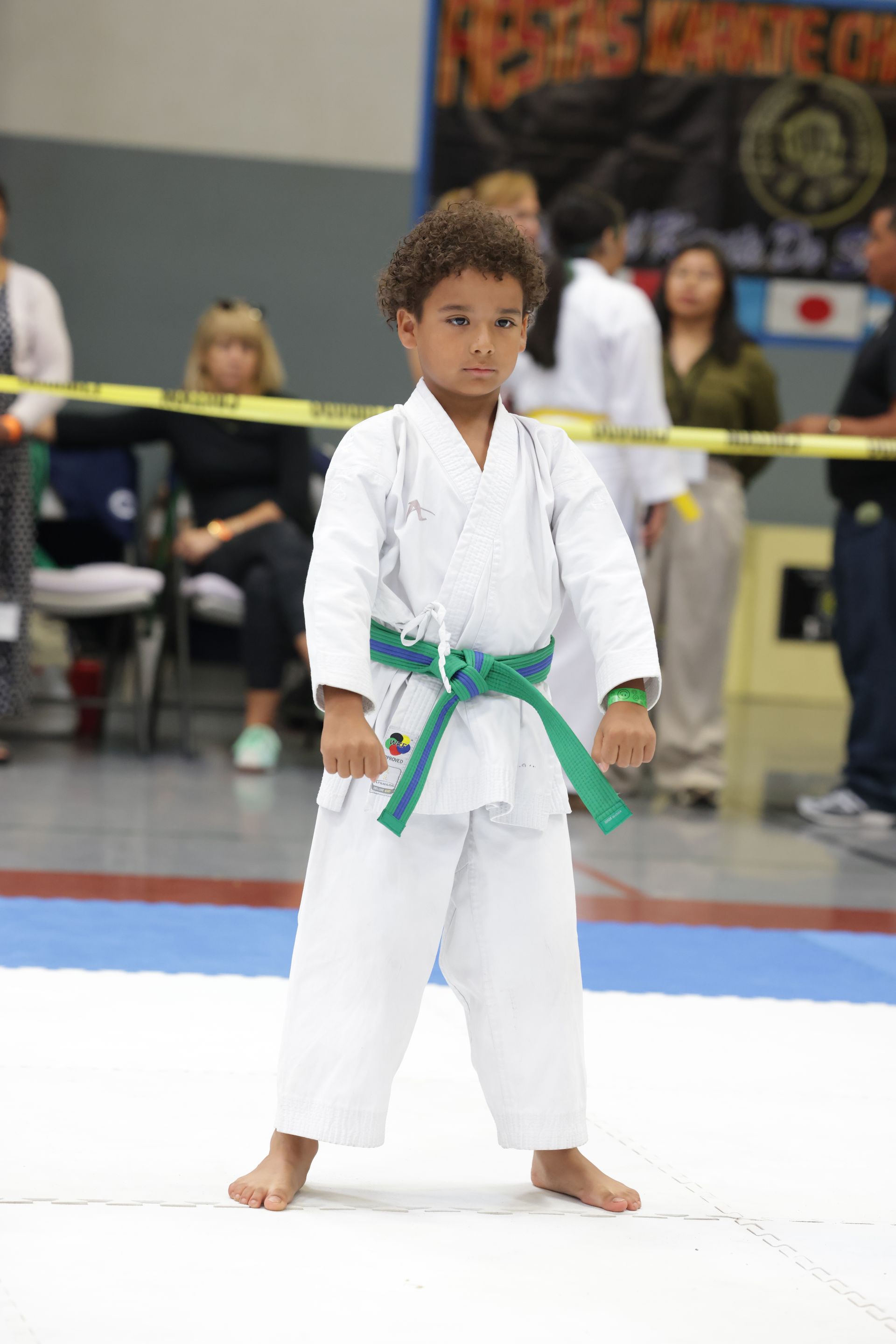 Child in a white karate gi with green belt standing on a mat at a martial arts competition.