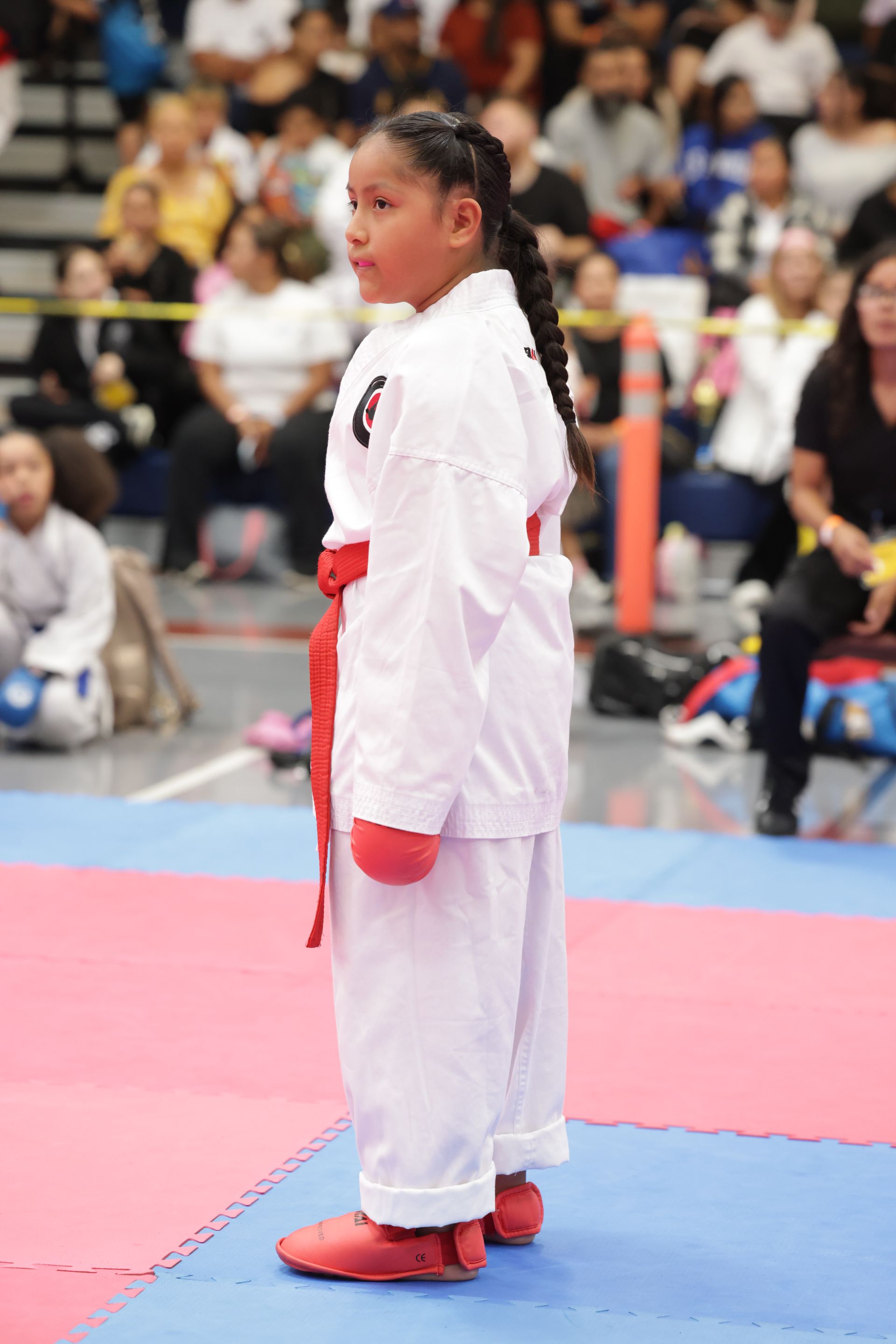 Martial artist in white uniform and red gloves standing on a blue mat at an indoor competition