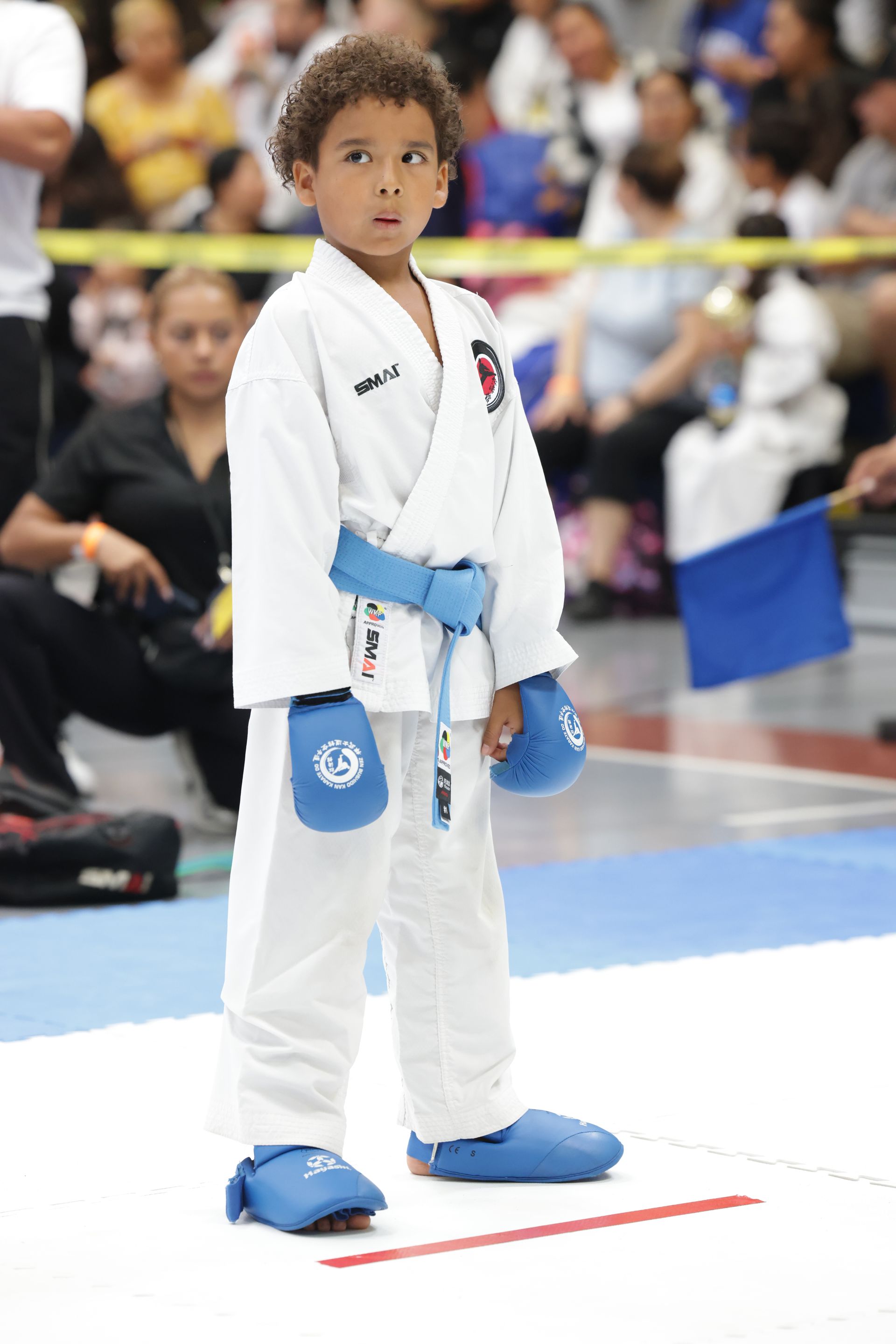 Young karate competitor in a white gi with blue gloves and belt stands on a mat at an indoor tournament.