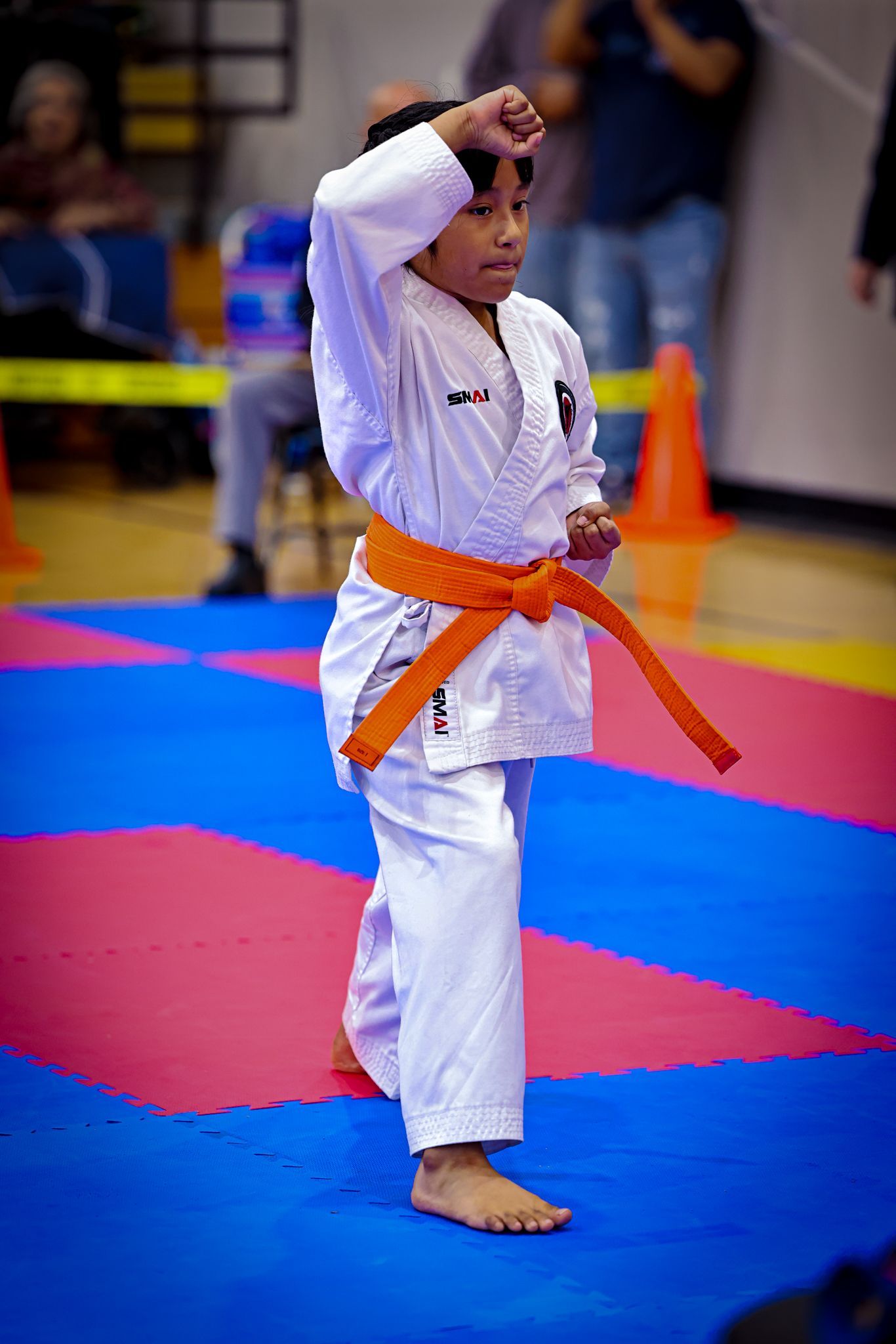 A person in a white karate uniform with an orange belt performing a high block on a blue and pink mat in a gym.