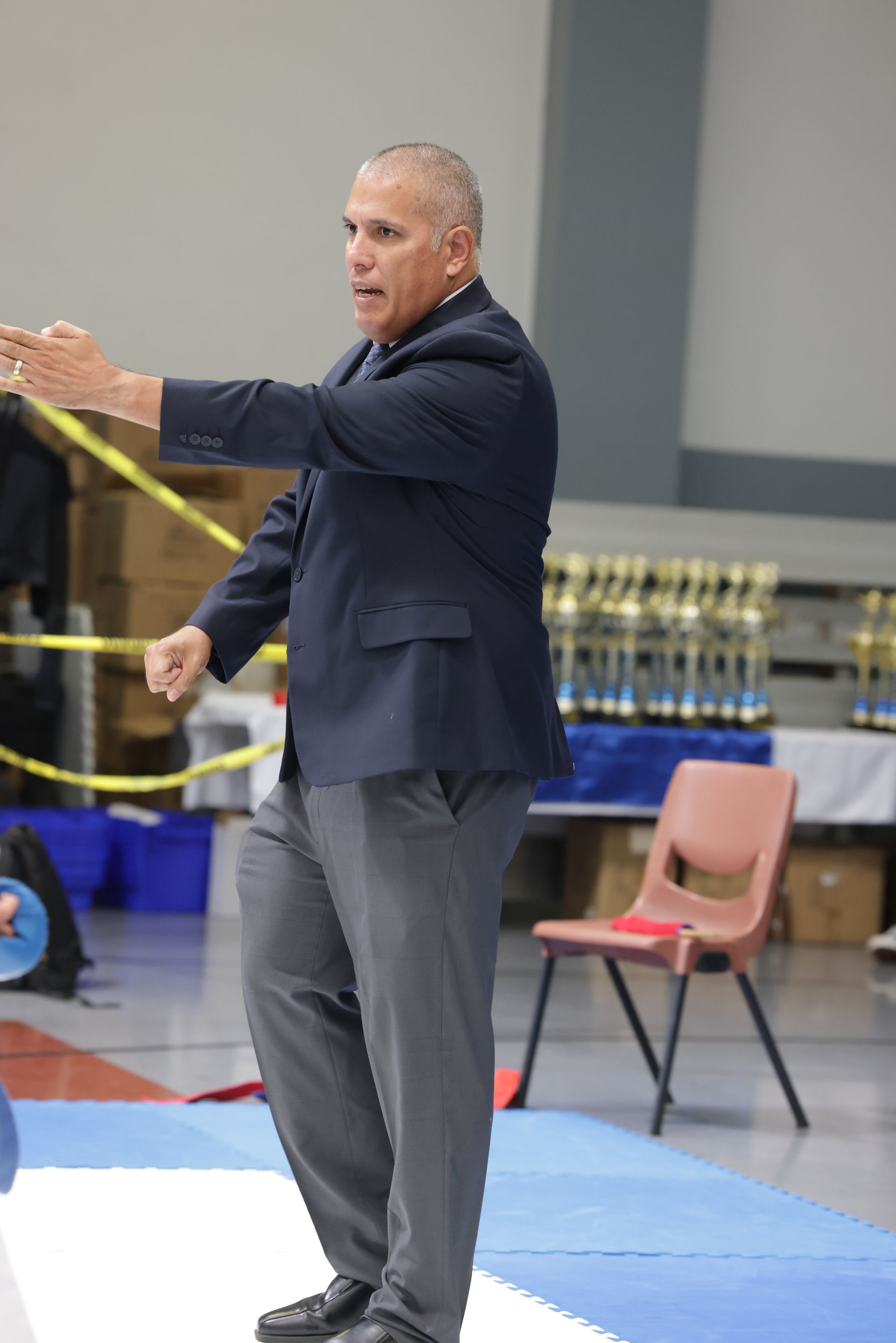 Man in a dark suit gesturing on a gym mat, with chairs and trophies in the background.