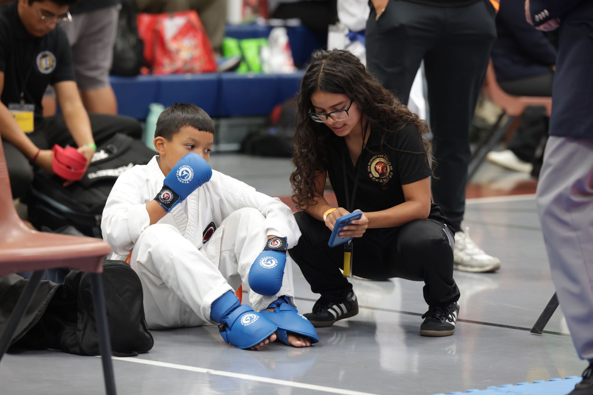 Martial arts match with a child in blue gloves kneeling as a coach assists on the mat