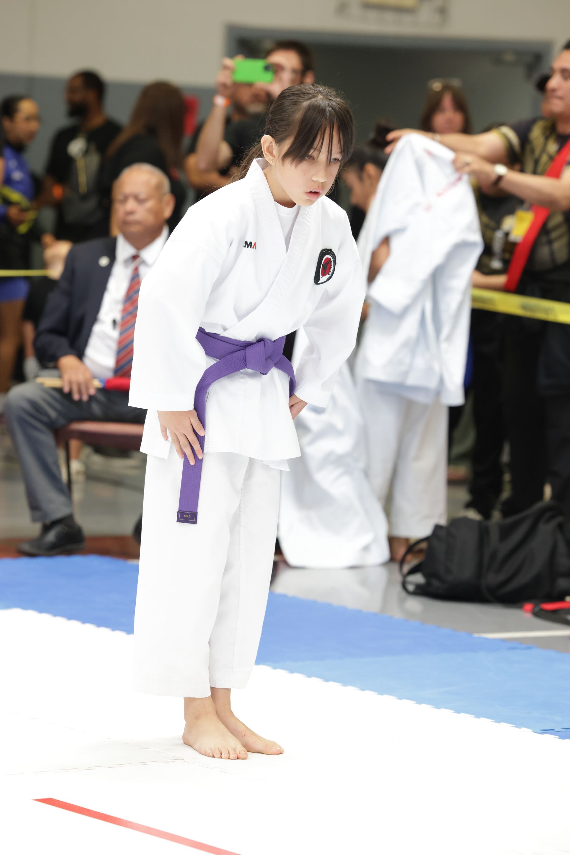 Martial artist in white gi with purple belt bows on a competition mat, spectators behind.