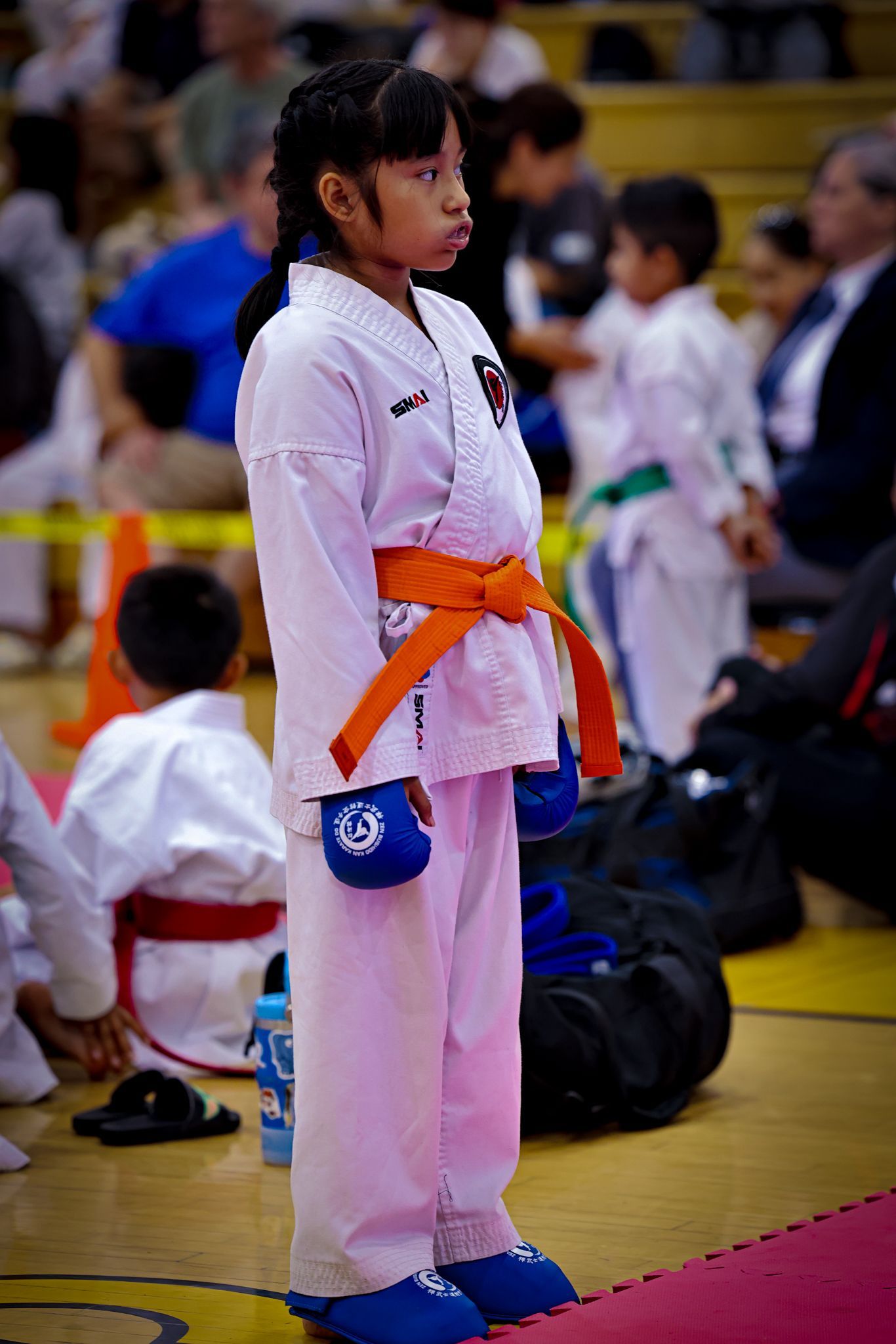 A child in a white karate uniform with an orange belt stands on a gym mat during a martial arts competition.