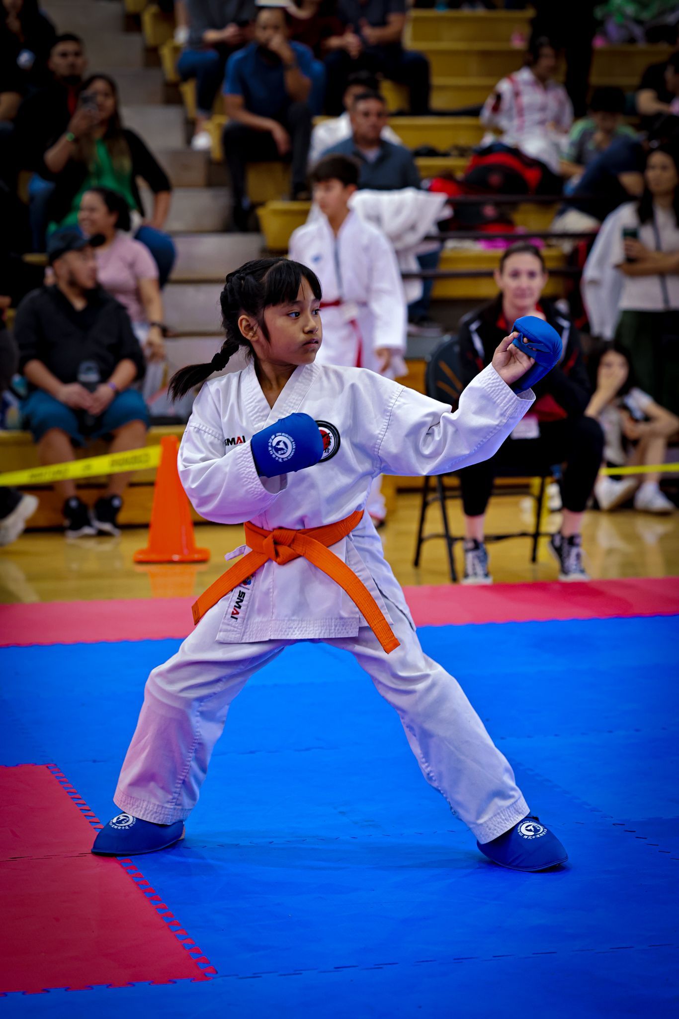 A martial artist in a white uniform and orange belt performs a punch while wearing blue sparring gloves on a blue mat.