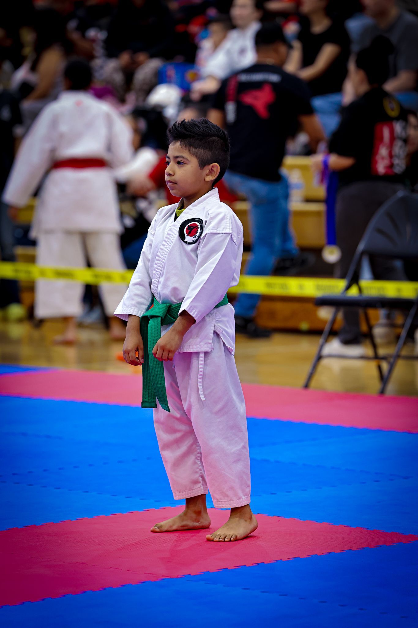 A child in a white karate uniform with a green belt stands barefoot on a blue and red mat during a tournament.