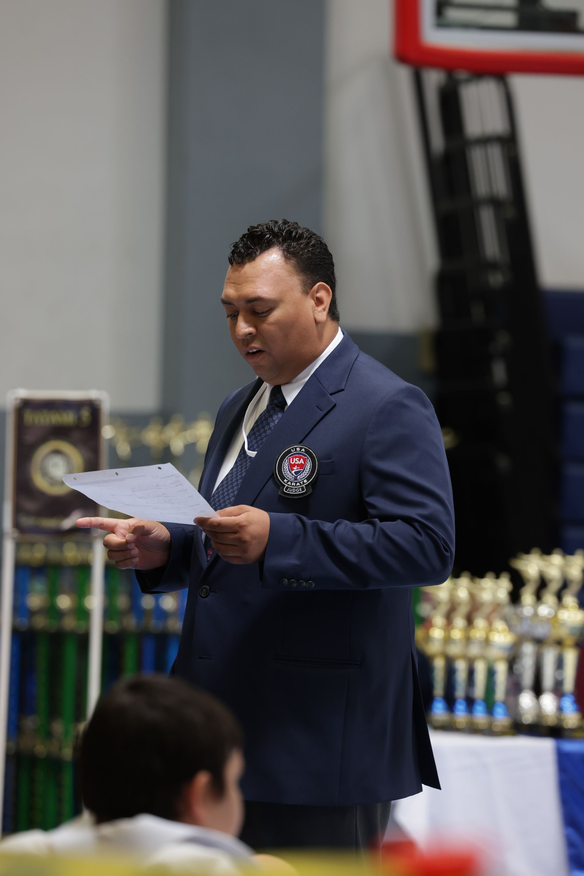 Man in a navy suit reading from papers at an indoor awards ceremony, with trophies on a table behind him