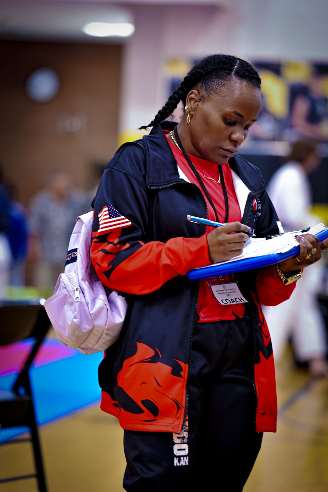 A person in a red and black jacket with an American flag patch writing on a blue clipboard, holding a light pink bag.