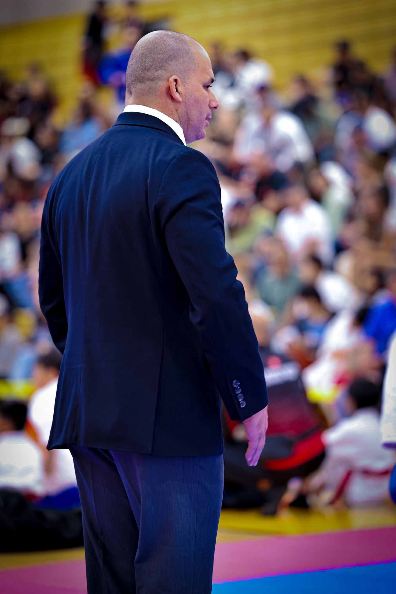 A person in a dark suit stands facing a blurred sports arena, observing a crowd seated on the floor.