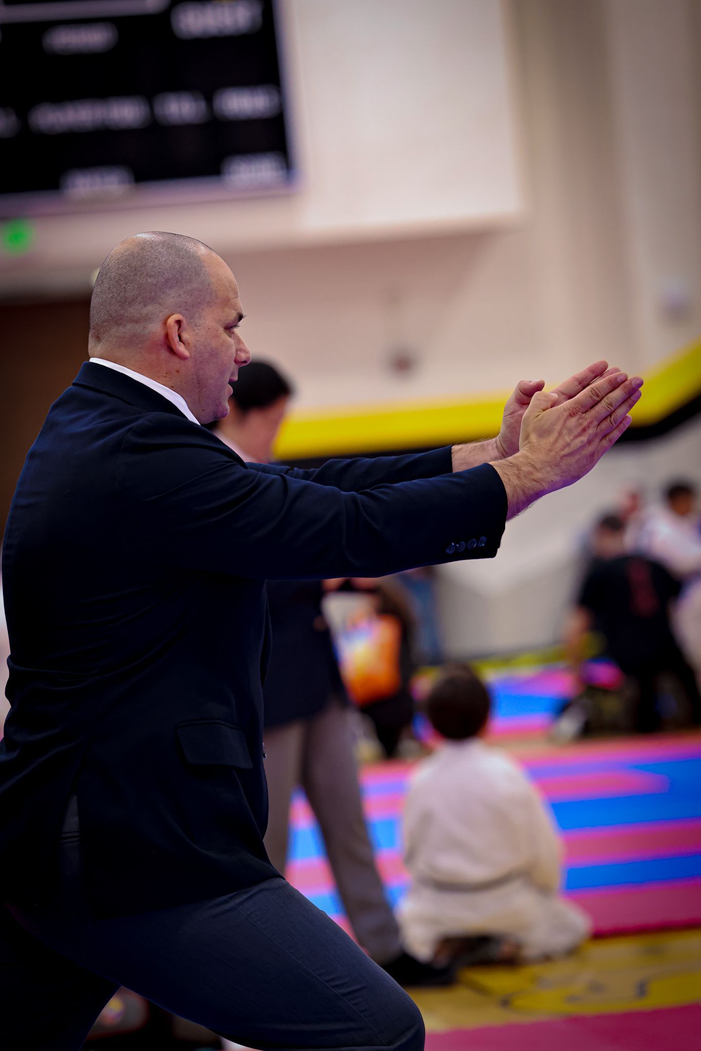 A person in a suit demonstrating a martial arts hand position at a tournament, with spectators in the background.