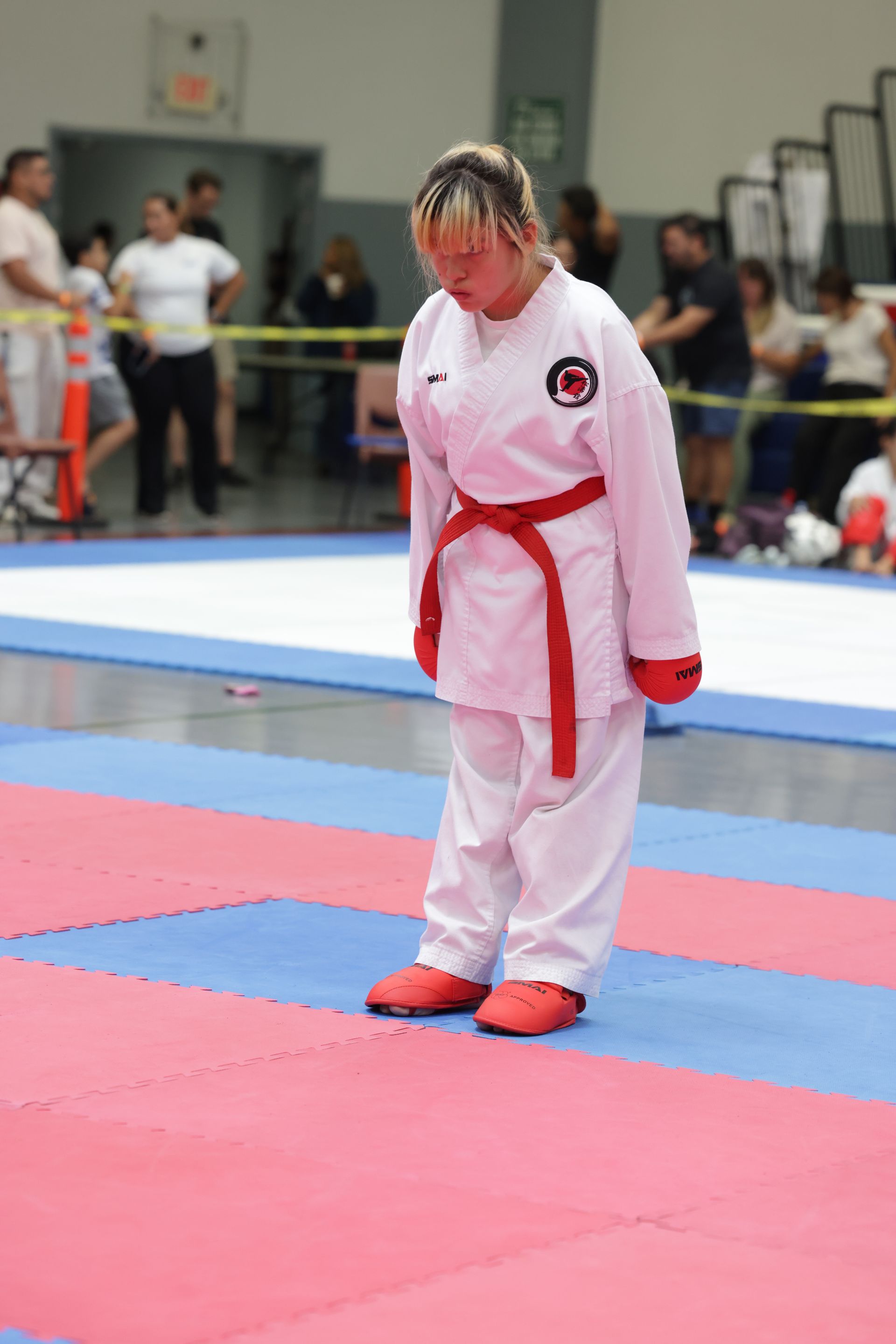 Martial artist in a white gi with red belt stands on a red-and-blue mat at a competition.