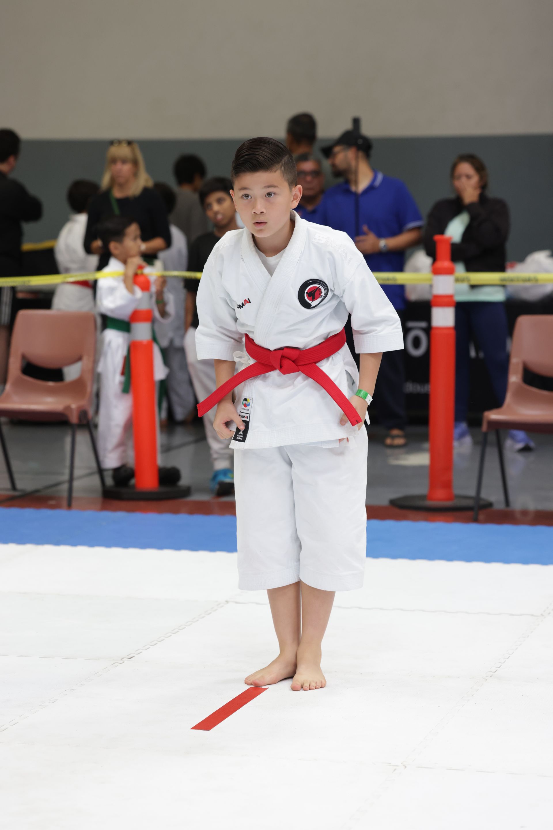Young martial artist in a white gi with red belt stands on a competition mat, with spectators behind him.